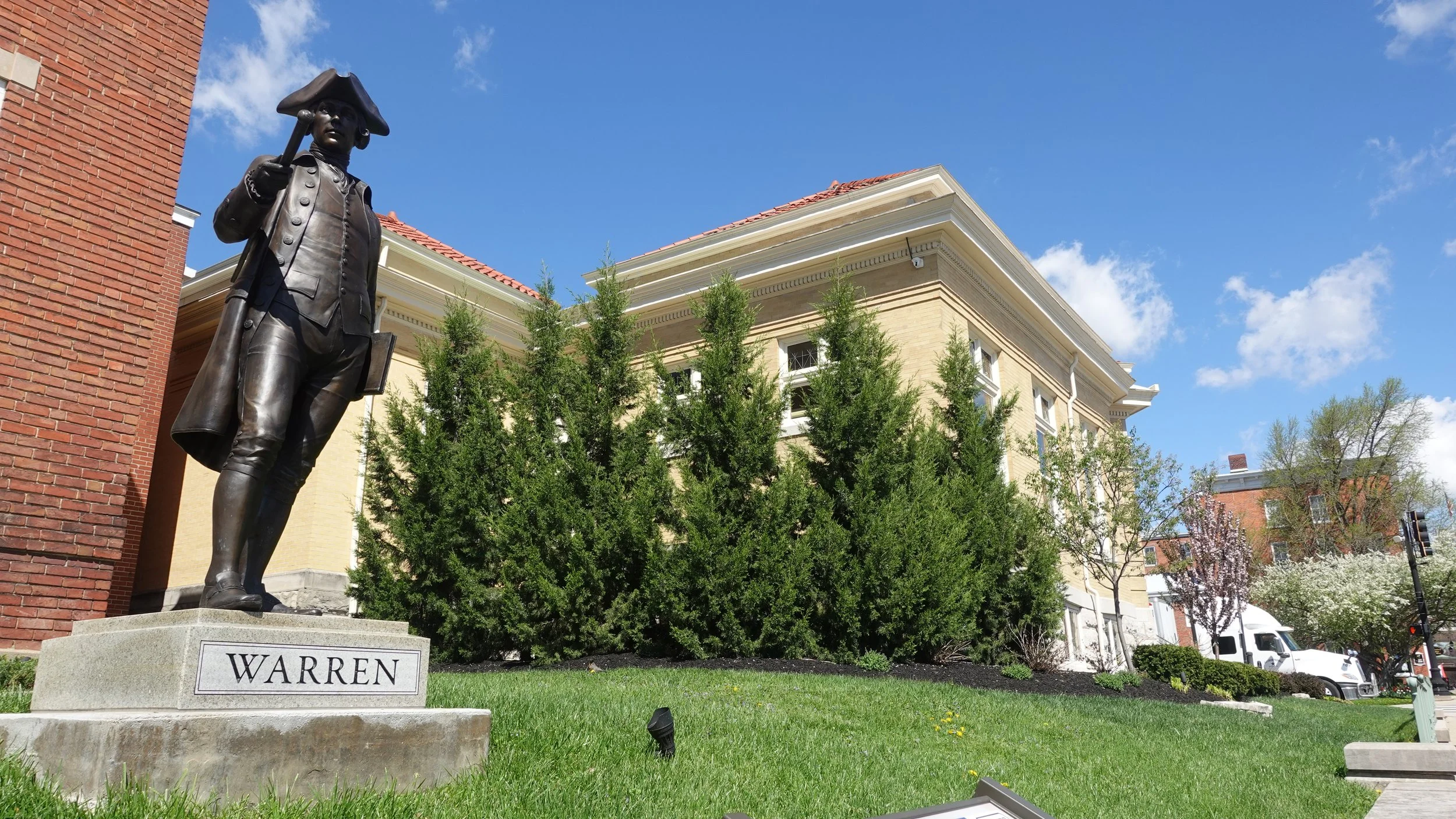 Bronze statue of a man in historical clothing, holding a hat in one hand, standing on a stone pedestal with a plaque labeled "WARREN". The statue is placed in front of trees and buildings with red-tiled roofs under a blue sky with some clouds.