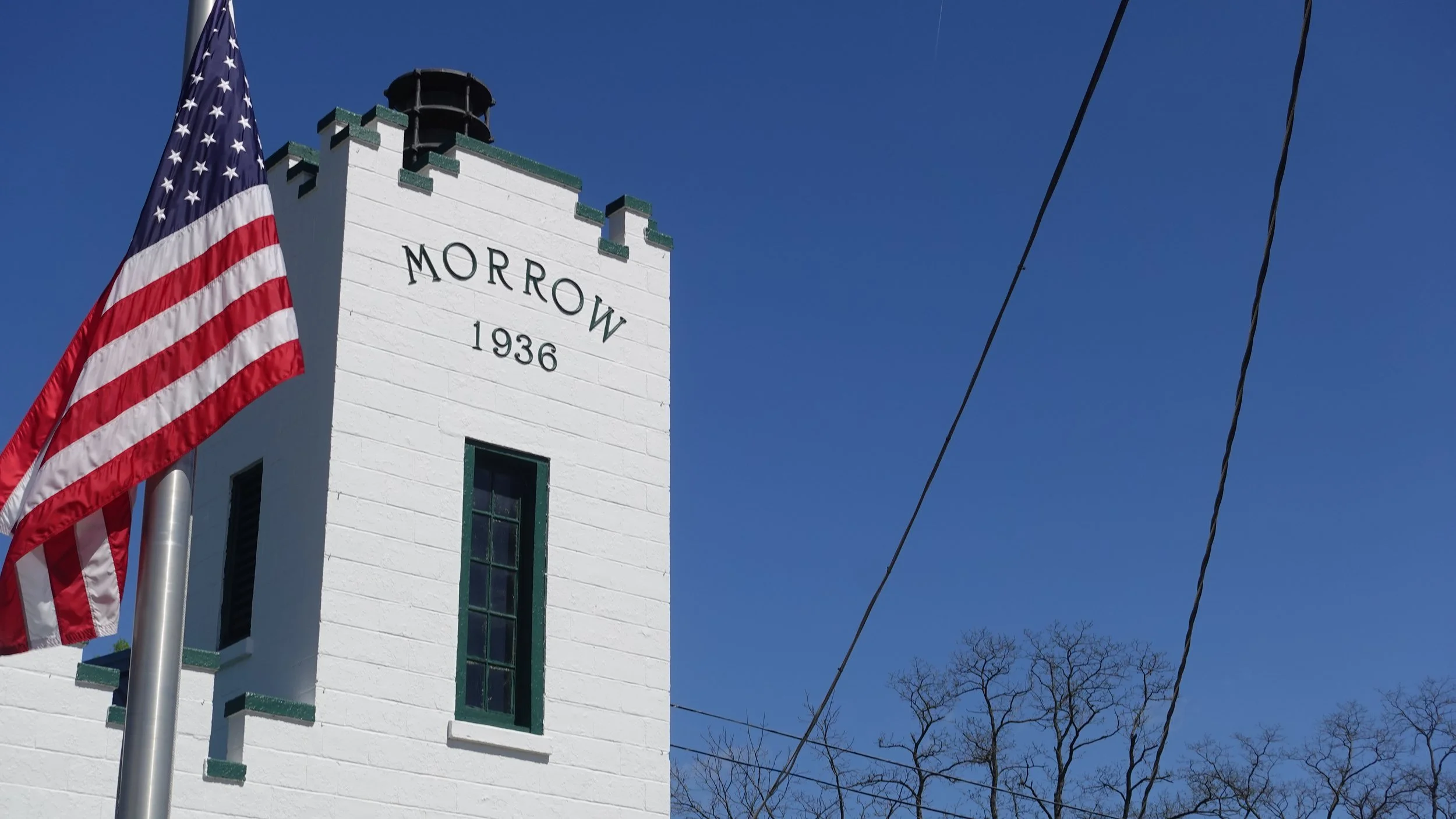 A white historic building with a tower displaying the words "MORROW 1936" on the side, an American flag flying in front, and a clear blue sky.