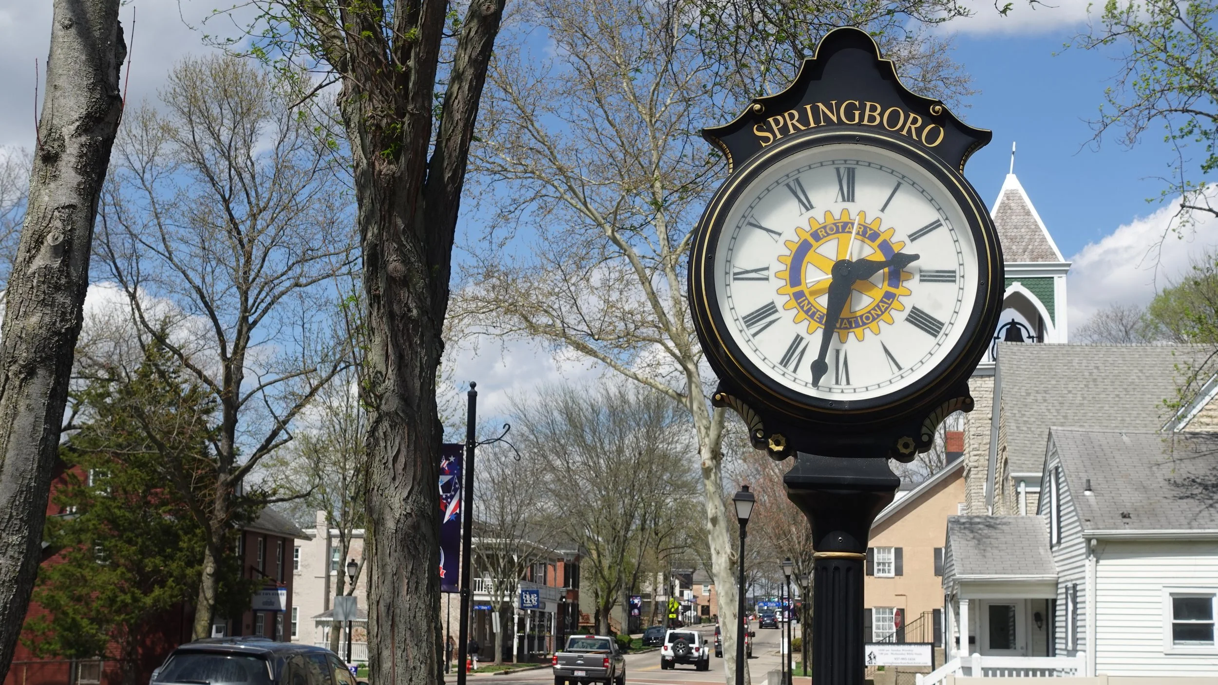 A street clock with the name 'Springboro' displayed at the top, surrounded by leafless and budding trees, with residential houses and colorful banners hanging along the street in the background.