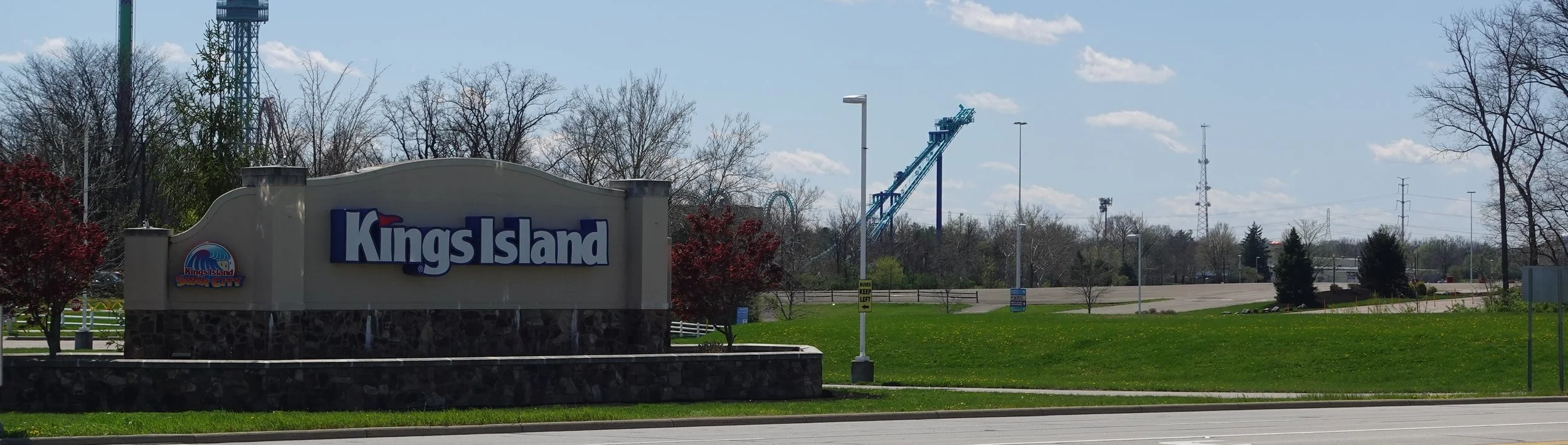 Entrance sign for Kings Island amusement park on a sunny day with trees, a roller coaster track, and parking lot in the background.