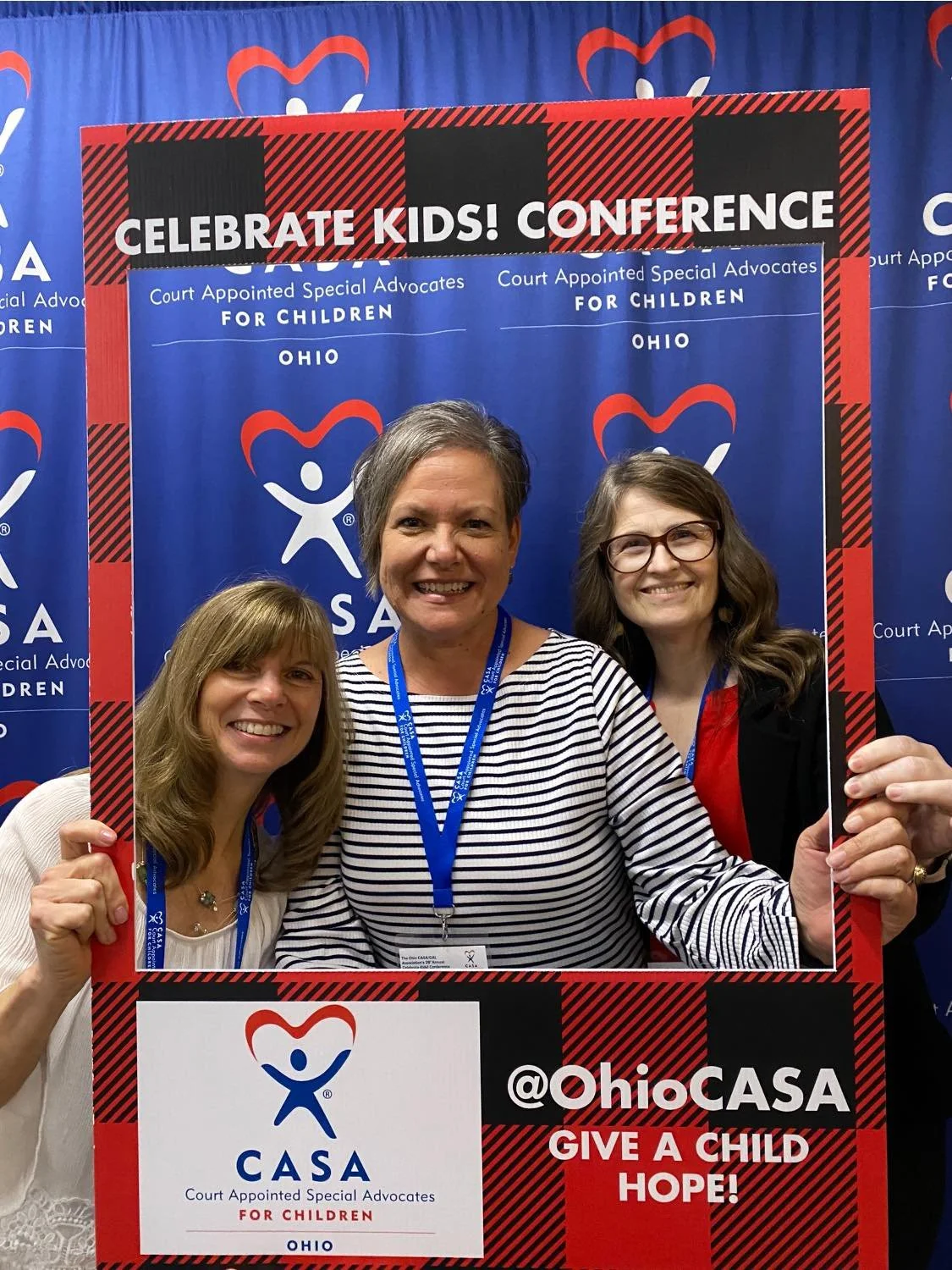 Three women posing behind a red and black frame at Ohio CASA's Celebrate Kids Conference, with a blue backdrop featuring CASA logos.