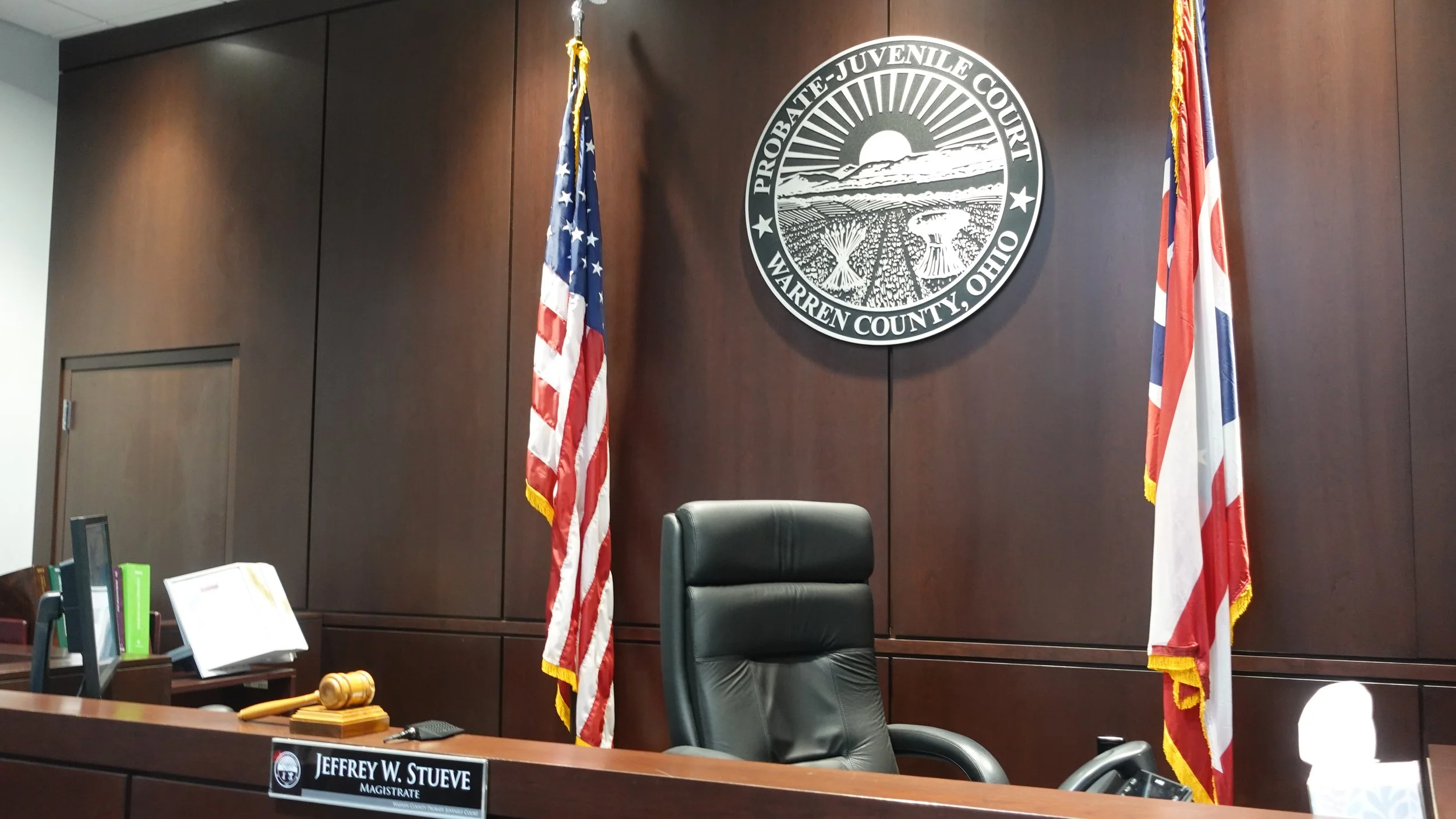 Inside a courtroom with a judge's bench, a leather chair, flags, a gavel, and a seal of Warren County, Ohio, on the wall.