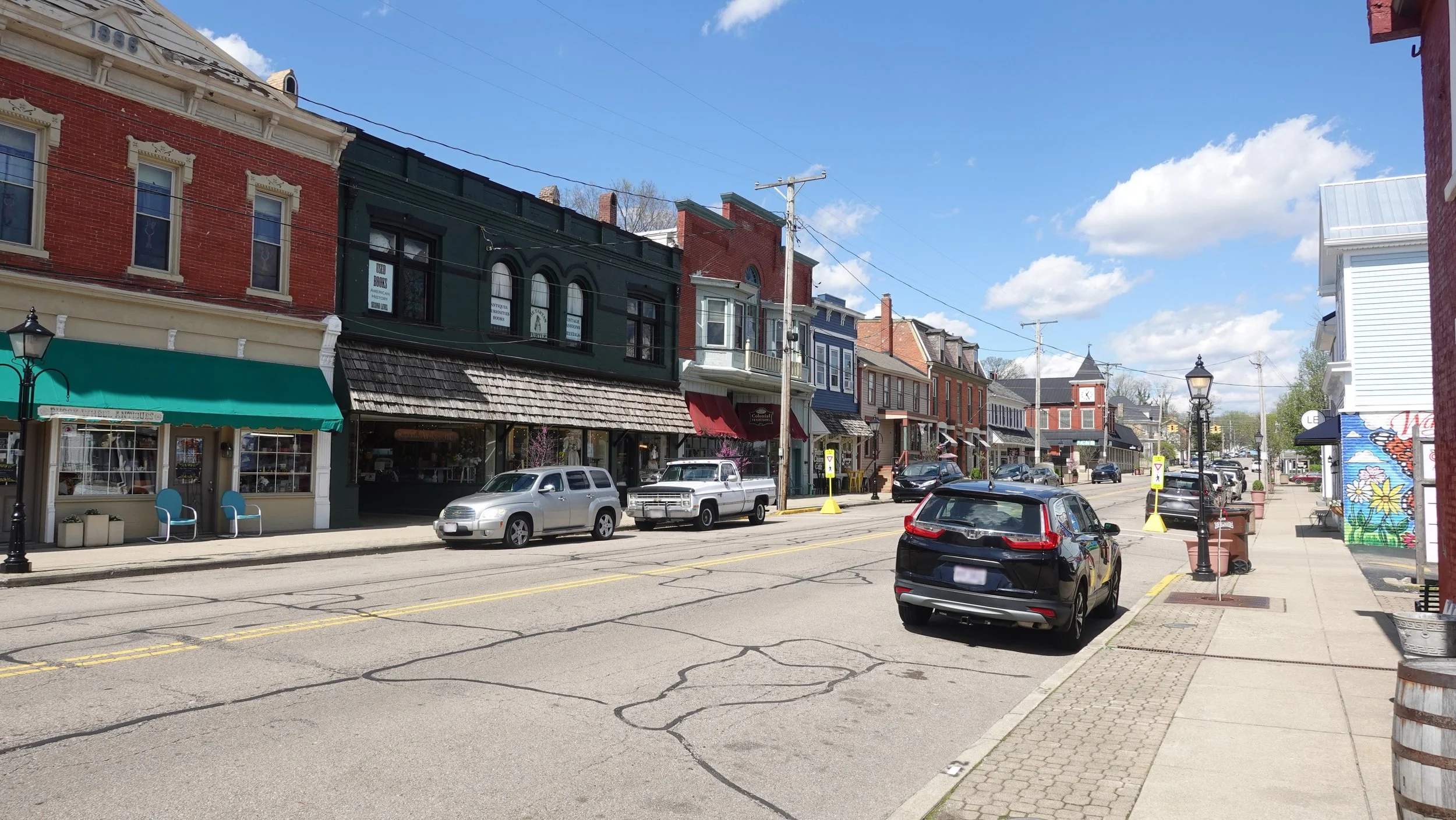 A small downtown street lined with colorful historic buildings, storefronts, parked cars, and a clear blue sky with scattered clouds.
