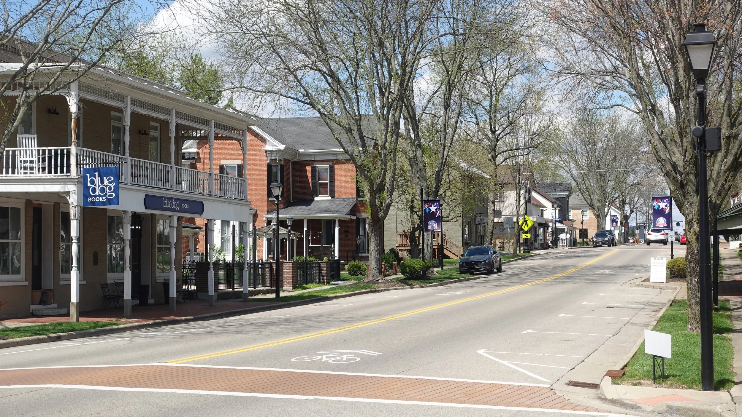 A small town street with trees, brick and wood buildings, street lamps, cars, and banners hanging on poles, in sunny weather with a blue sky and some clouds.
