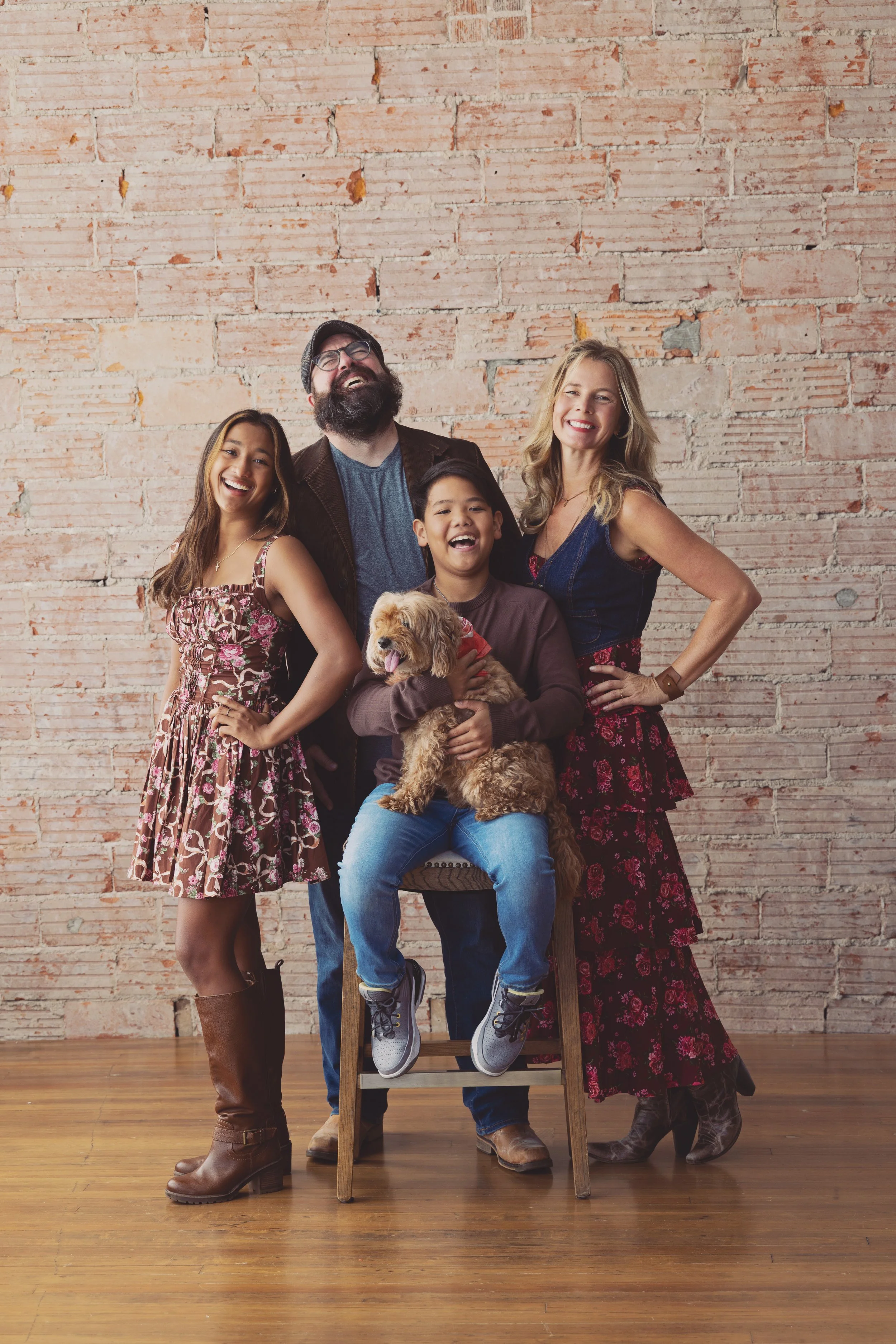 A diverse group of five people, including two women, two boys, and a man, are standing and sitting against a brick wall, smiling. One girl is wearing a floral dress with brown boots, the man has a beard and glasses, the boy in the middle is holding a small dog, and the woman has blonde hair and is wearing a floral skirt.