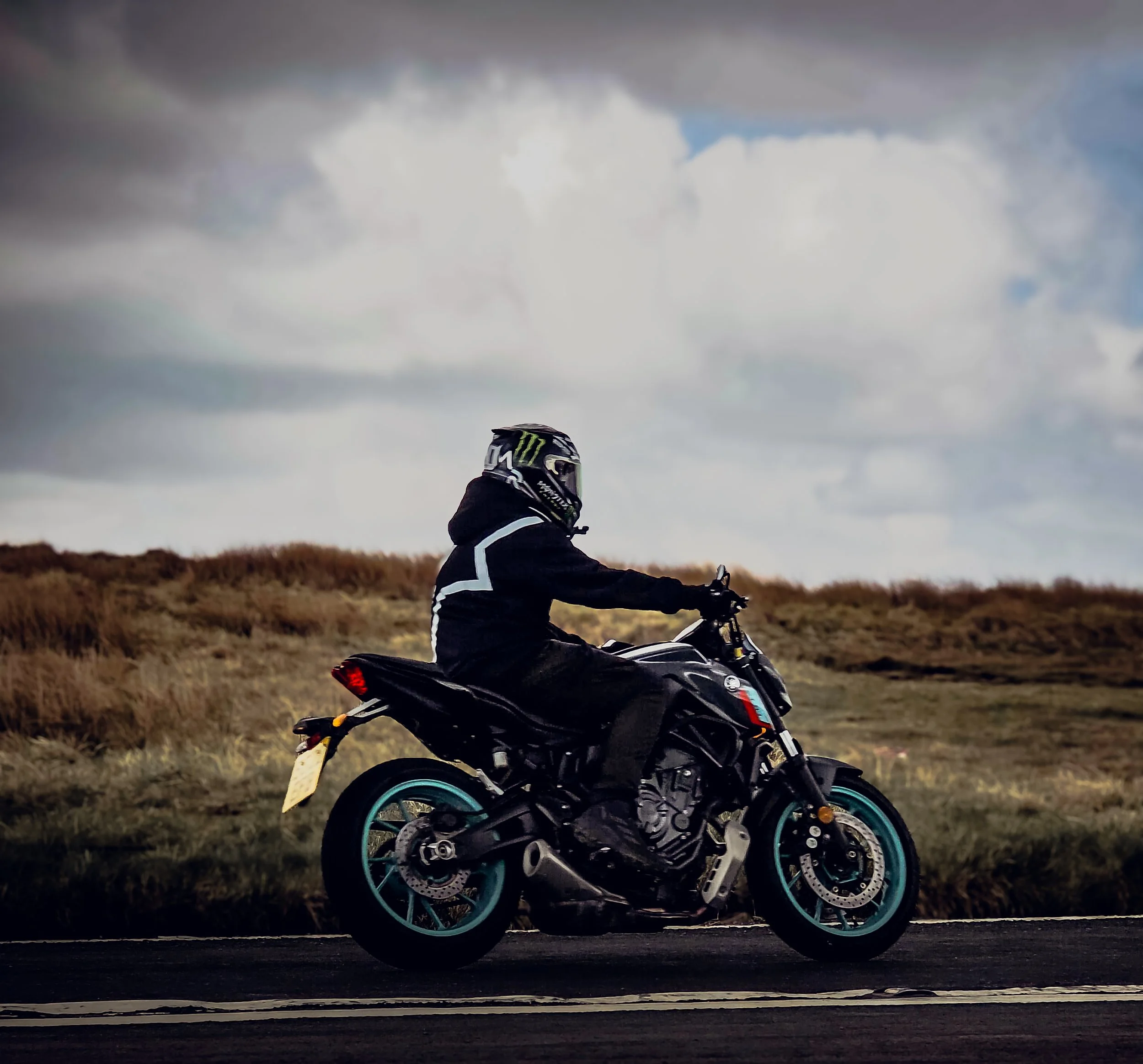 Motorcycle rider sitting on a black motorcycle in an open field under a cloudy sky.