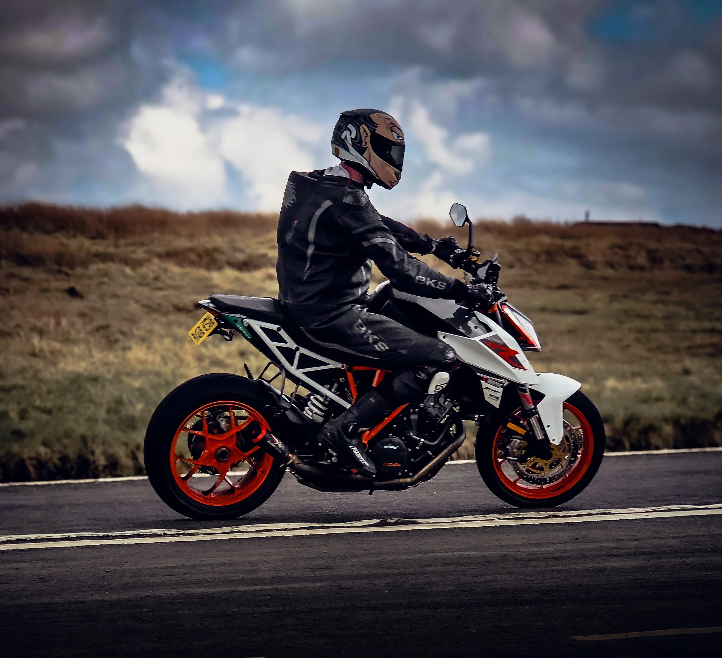 A motorcyclist wearing a black helmet and jacket riding a white motorcycle with orange wheels on a road with a cloudy sky in the background.