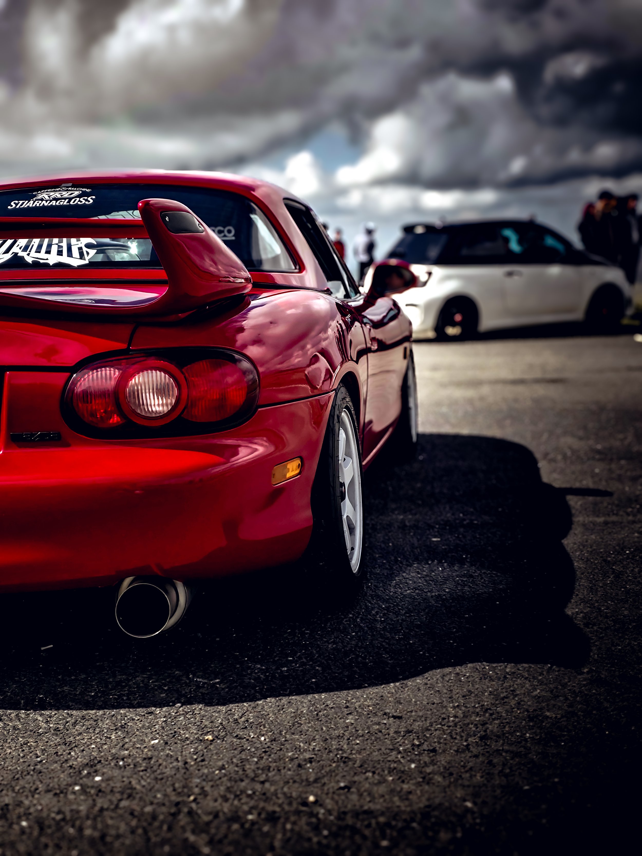 A red sports car parked on a dark pavement with a white car and people in the background under a cloudy sky.