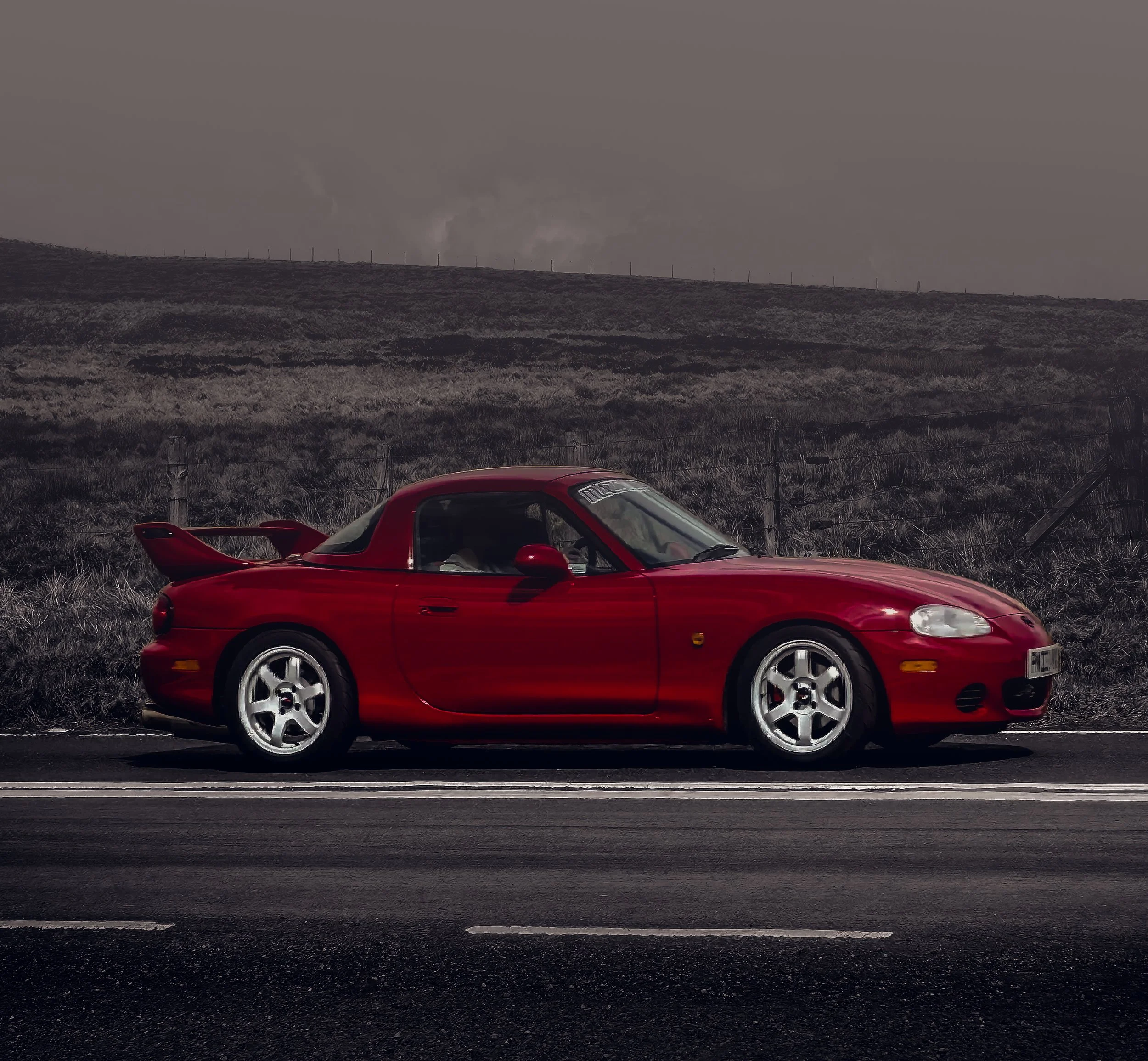 A red sports car driving on a paved road with grass and shrubs on either side. A hill with a fence and wind turbines is visible in the background under a cloudy sky.