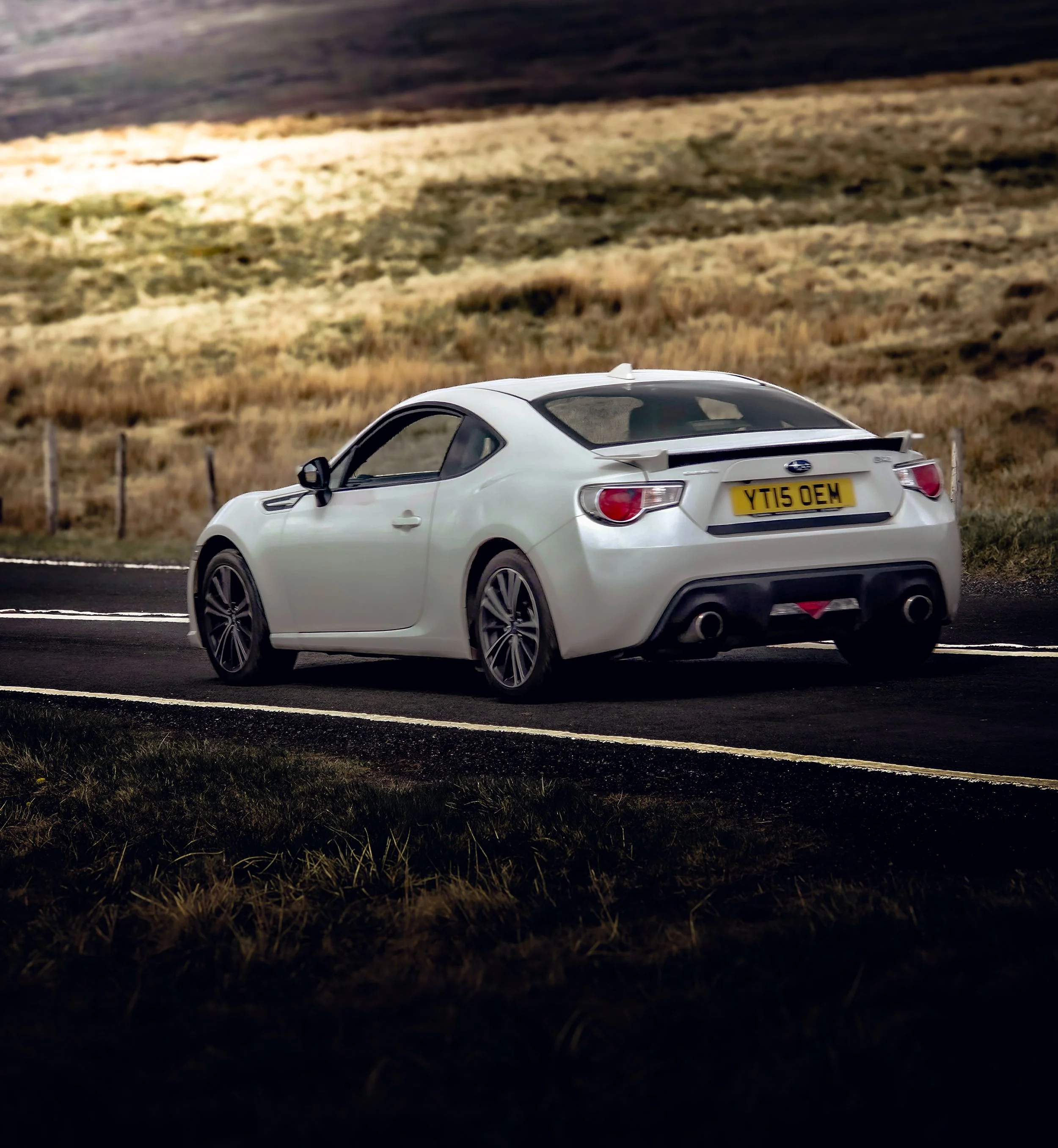White sports car driving on a rural road with grassy fields and hills in the background.