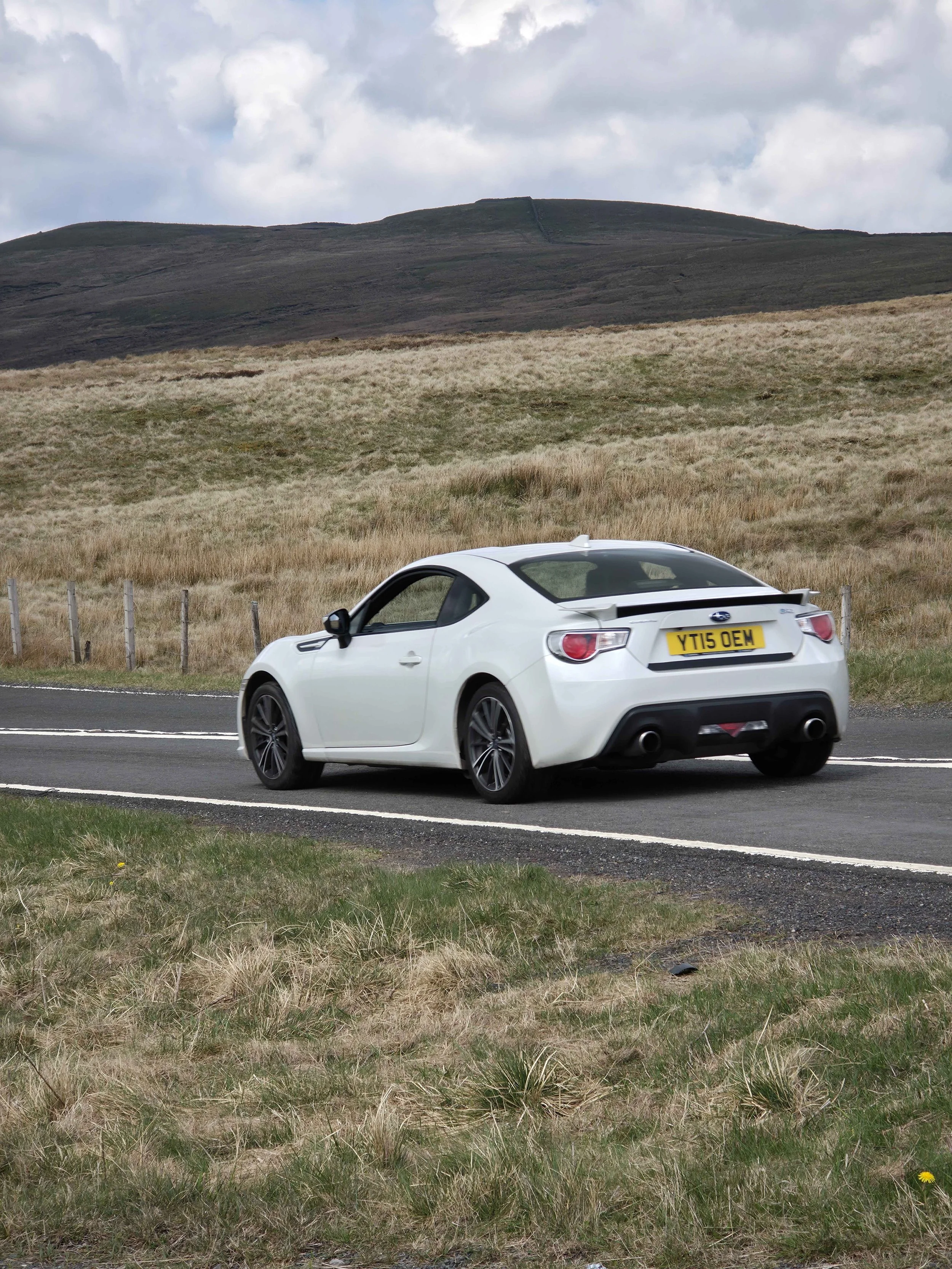 A white Subaru car with a UK license plate YT15 OEM driving on a road beside grassy fields with a hill in the background and a cloudy sky.