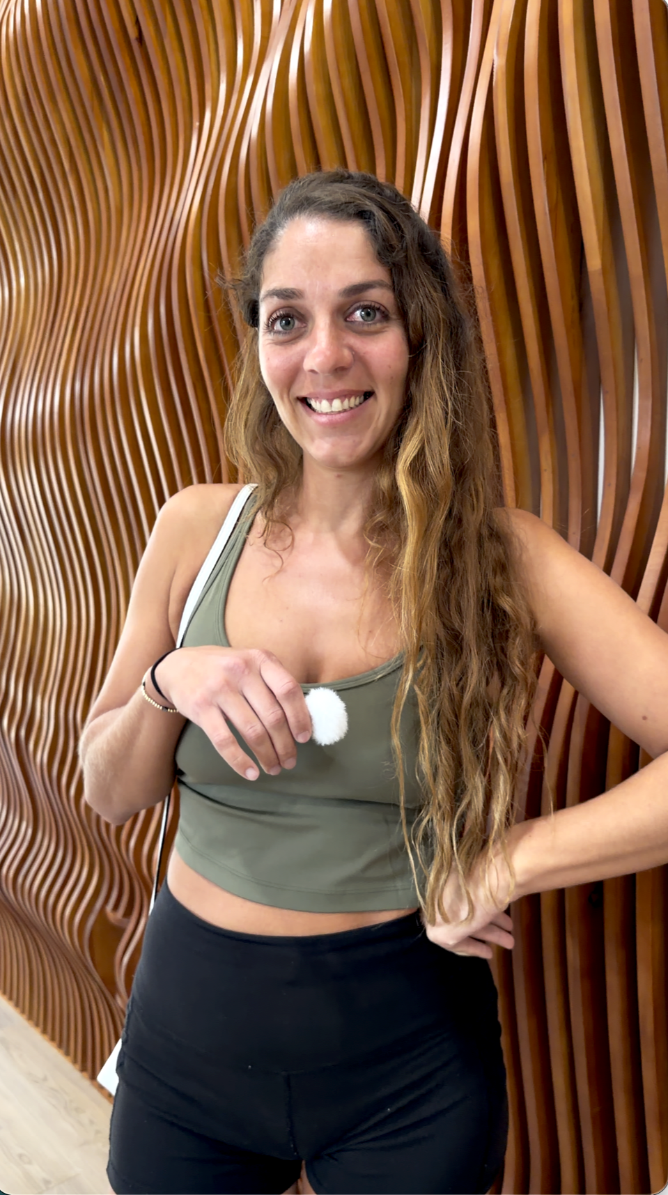 A young woman with long wavy hair, wearing a green sports bra and black leggings, smiling and standing in front of a wooden decorative wall.