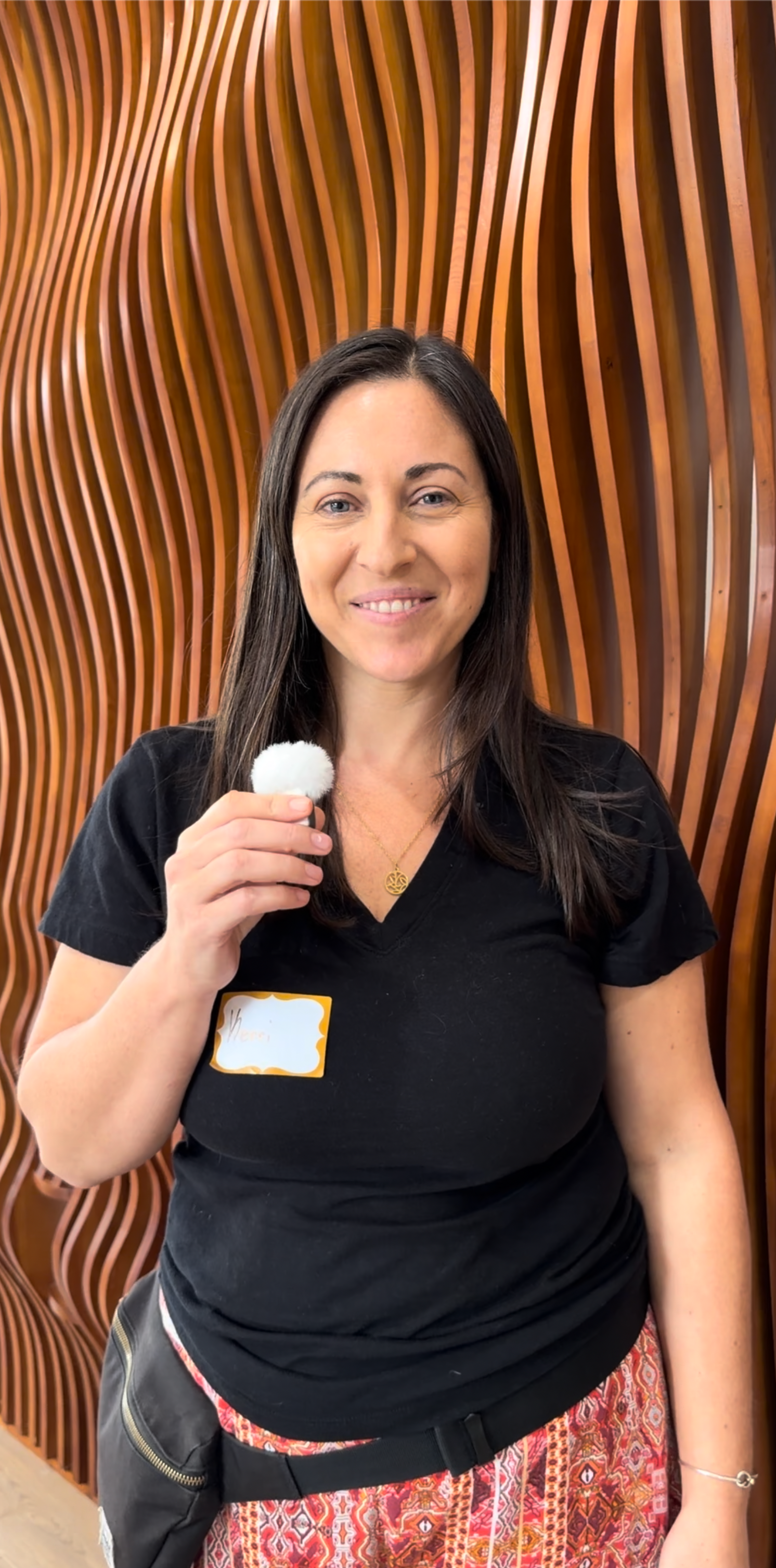 Woman with long dark hair holding a small white fluffy object. She is wearing a black T-shirt with a name tag, patterned skirt, and a gold necklace, standing in front of a wooden decorative wall.