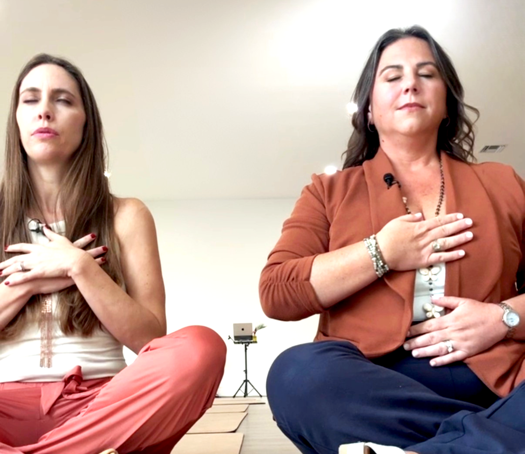 Two women seated with eyes closed and hands placed over their chests, in a meditative pose, in a bright indoor setting.