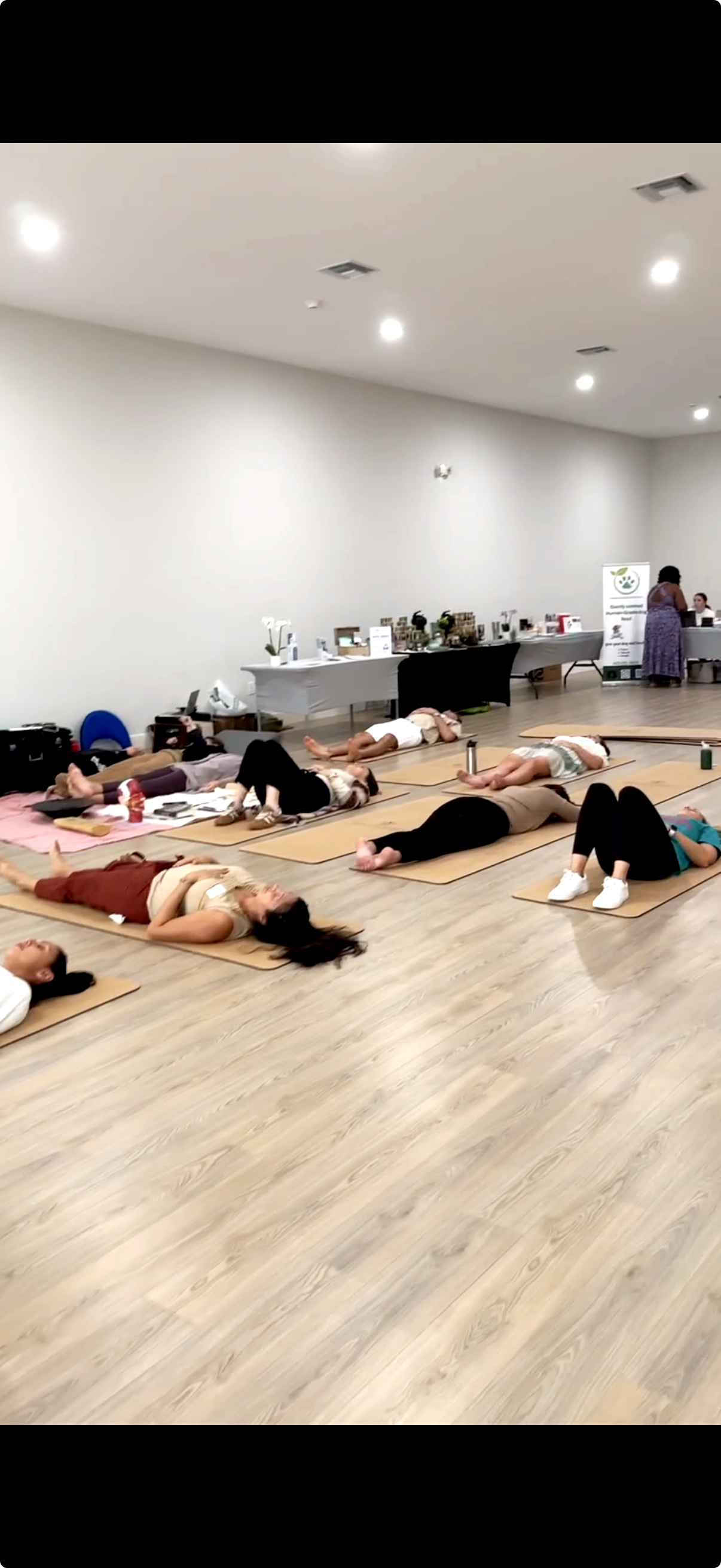 People participating in a yoga class, lying on mats in a spacious, well-lit room with a wood floor, while a few individuals are at a table in the background.