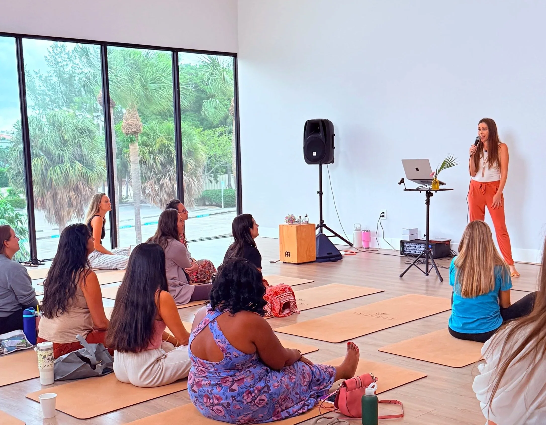A woman giving a presentation or speech standing next to a laptop on a stand with a microphone, inside a brightly lit room with large windows showing palm trees outside. The audience sits on mats on the floor, listening attentively.