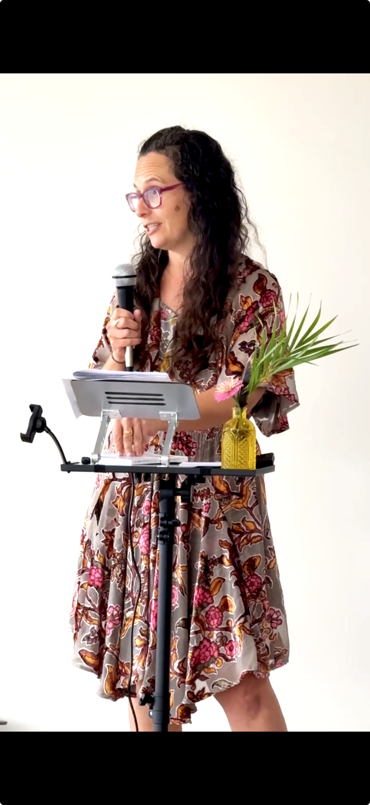 A woman with long curly dark hair, pink glasses, wearing a floral dress, standing at a music stand, holding a microphone, with a yellow vase containing a palm leaf and pink flower on the stand.