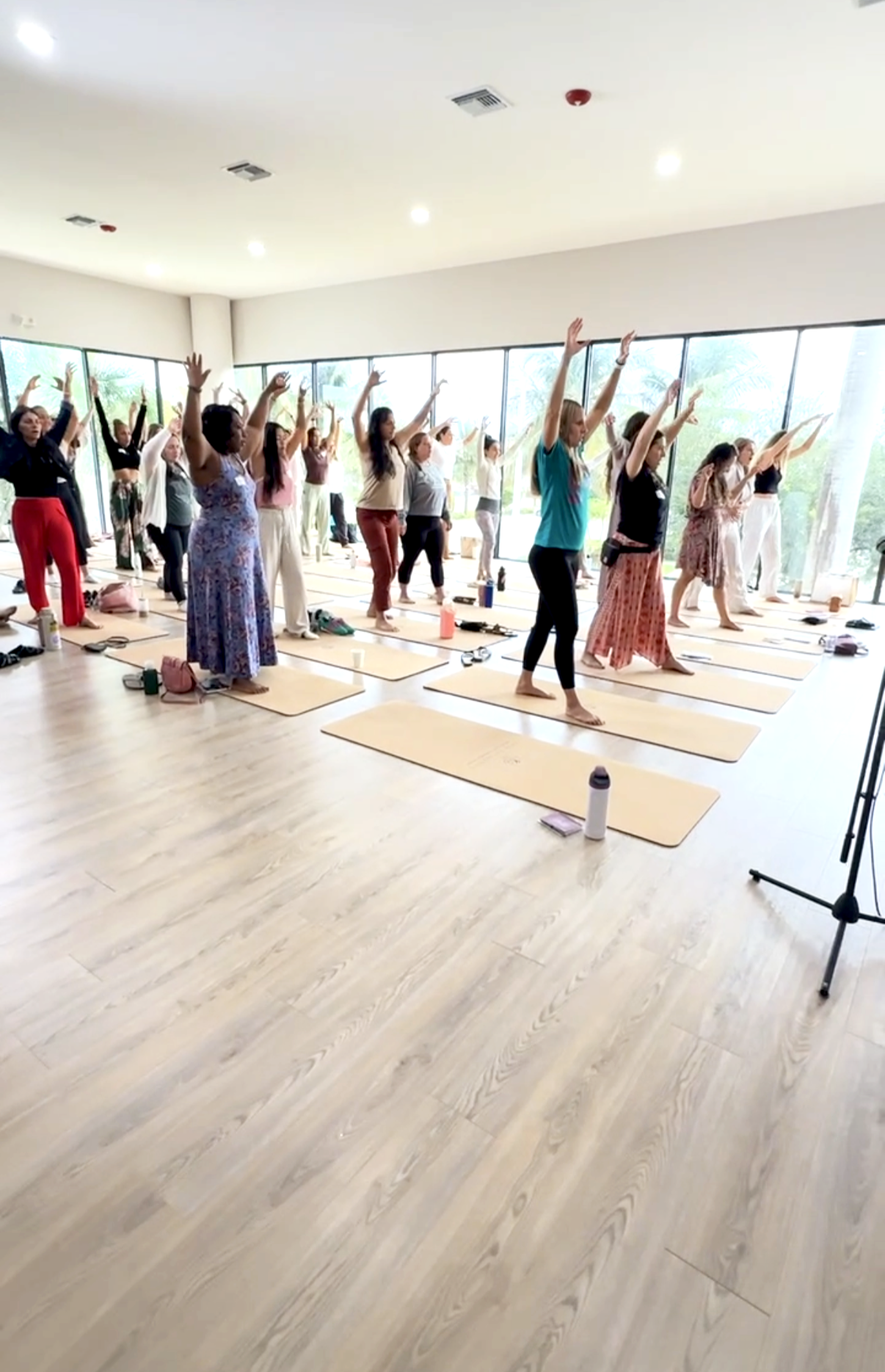 Group of people participating in a yoga class in a bright room with large windows, yoga mats, and water bottles.