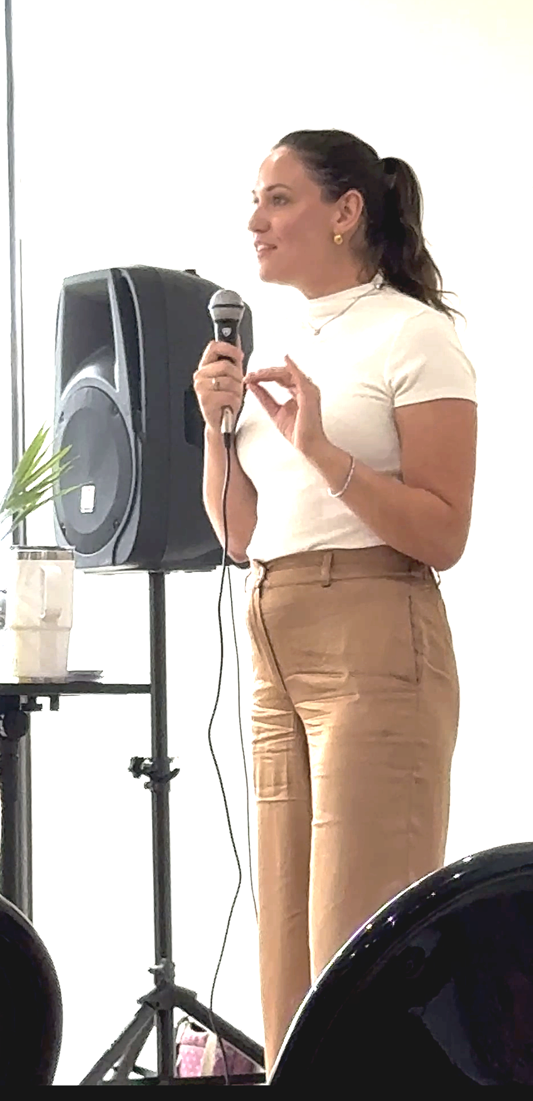 Woman speaking into a microphone near speaker, wearing a white top and beige pants, standing indoors with a plant in a pot nearby.