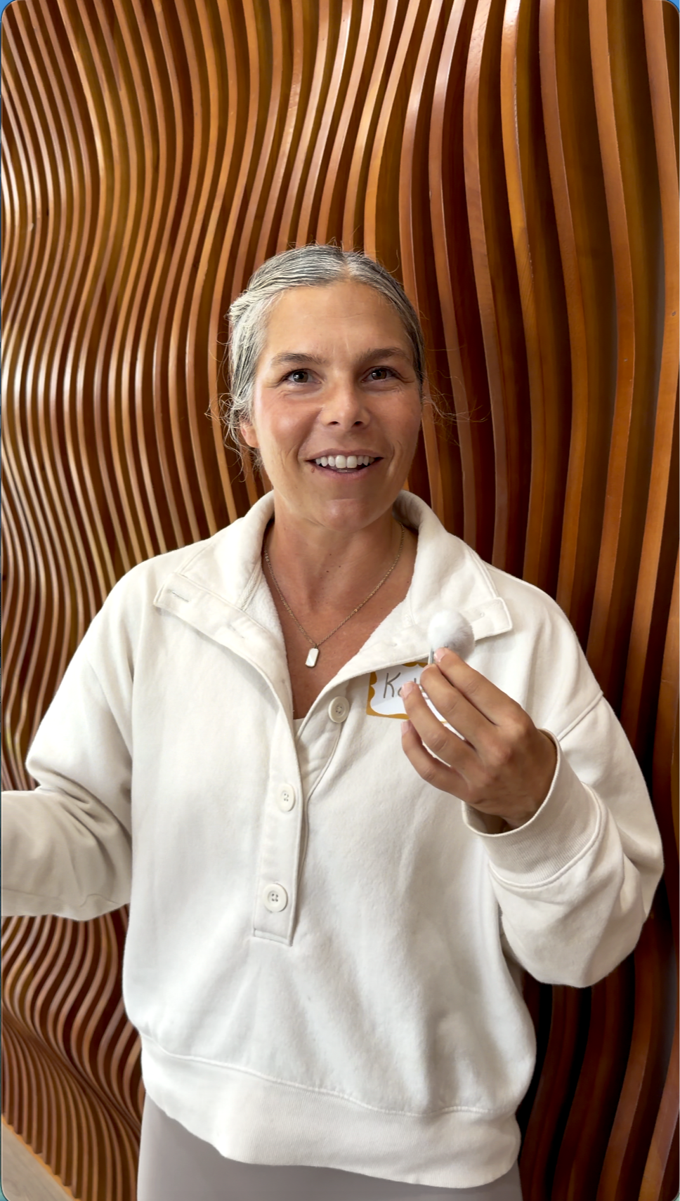 A woman with gray hair in a bun, wearing a cream-colored hoodie, holding a cotton ball, smiling, standing in front of a wooden wavy decorative wall.