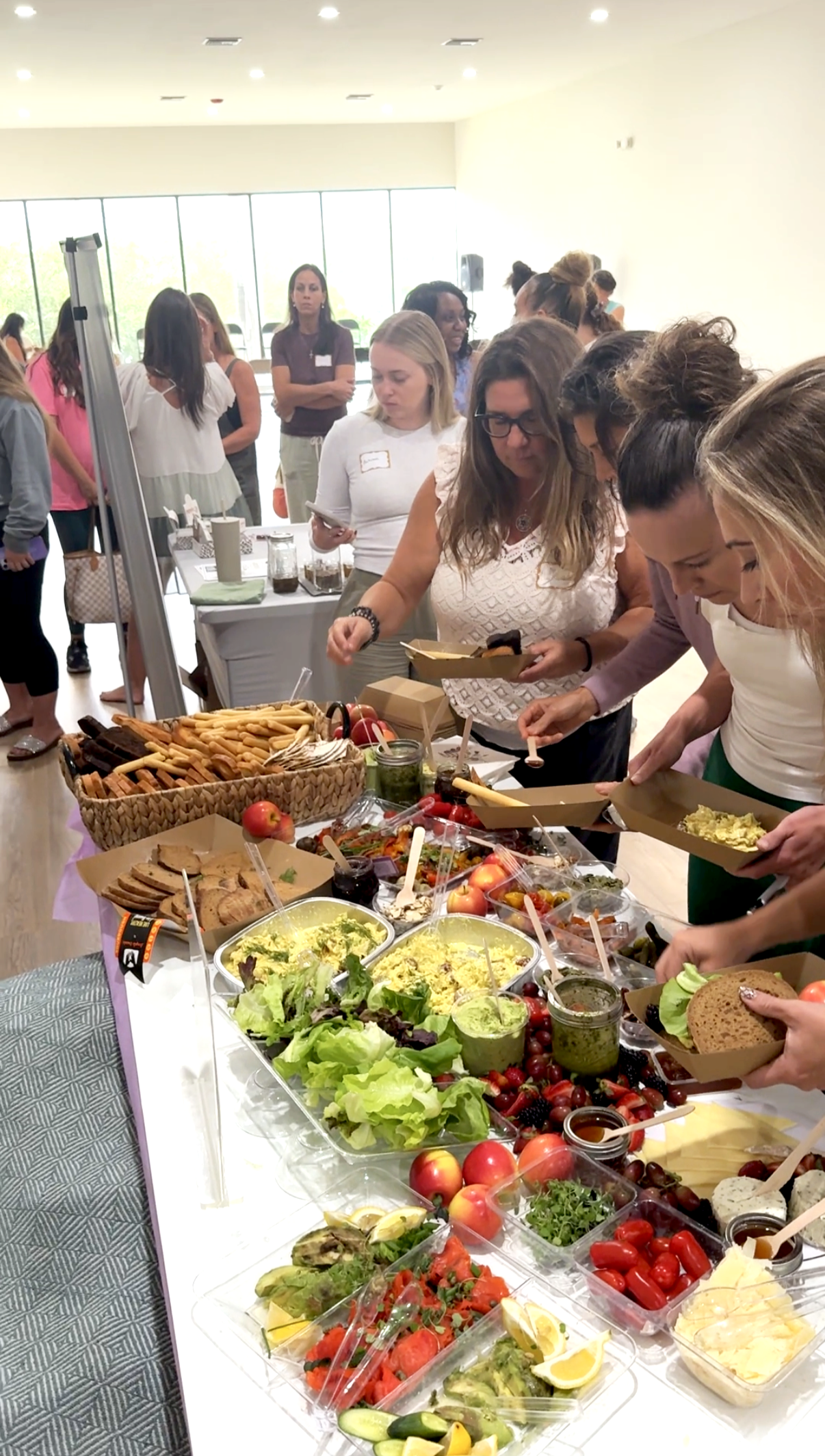 People serving themselves food at a buffet with salads, vegetables, bread, and various dishes in a bright room with large windows.