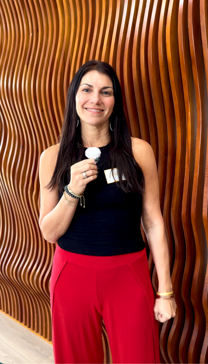 A woman with long dark hair wearing a black sleeveless top and red pants, smiling, holding a small microphone, standing in front of a wavy wooden wall.