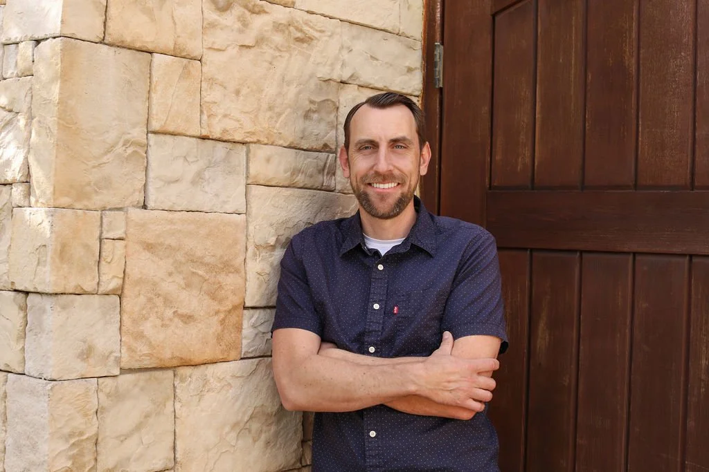 Man with dark short hair and beard smiling, standing next to a stone wall and a wooden door, arms crossed.