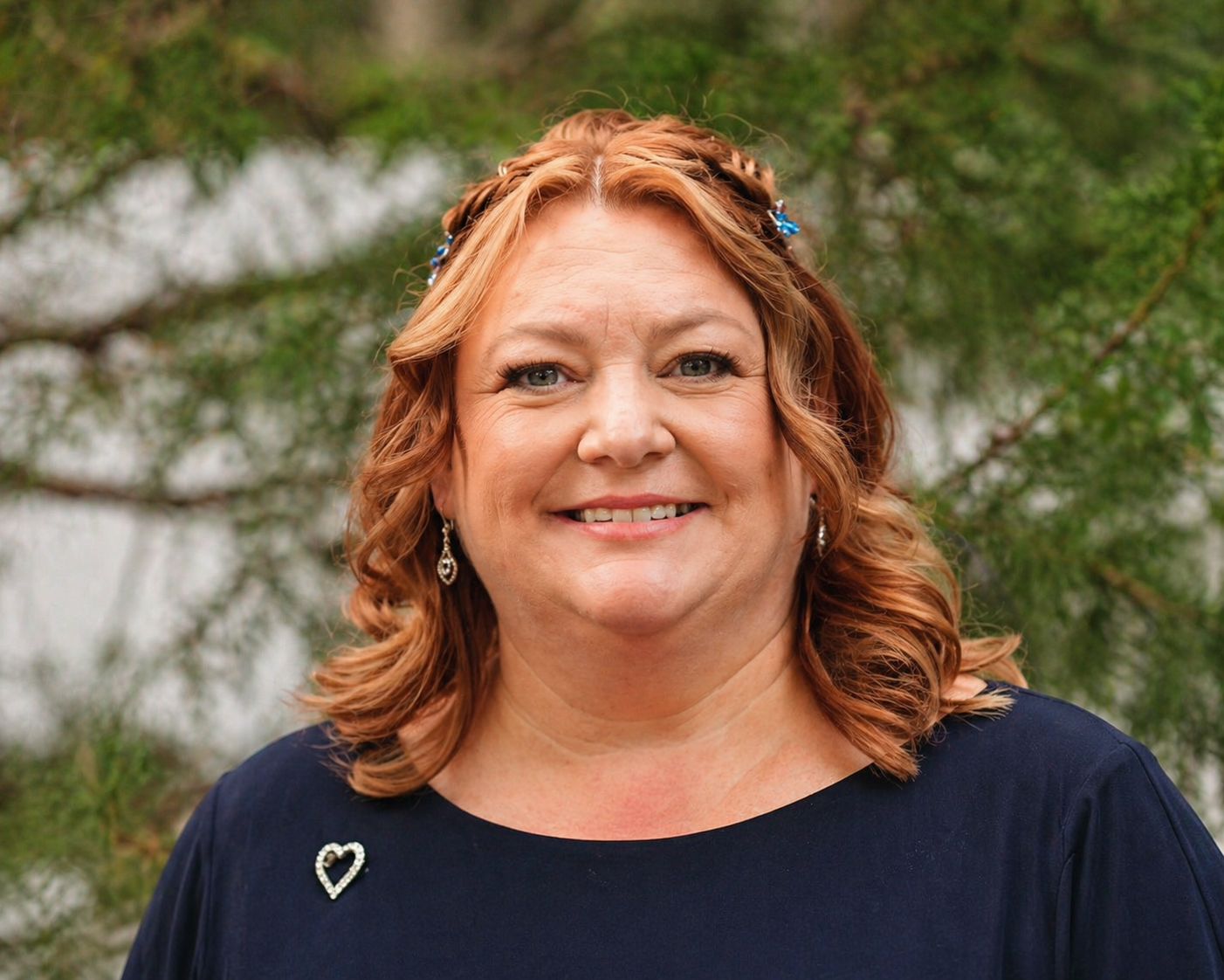 A smiling woman with curly red hair wearing earrings, a navy blue top with a heart-shaped pin, standing outdoors with green foliage in the background.