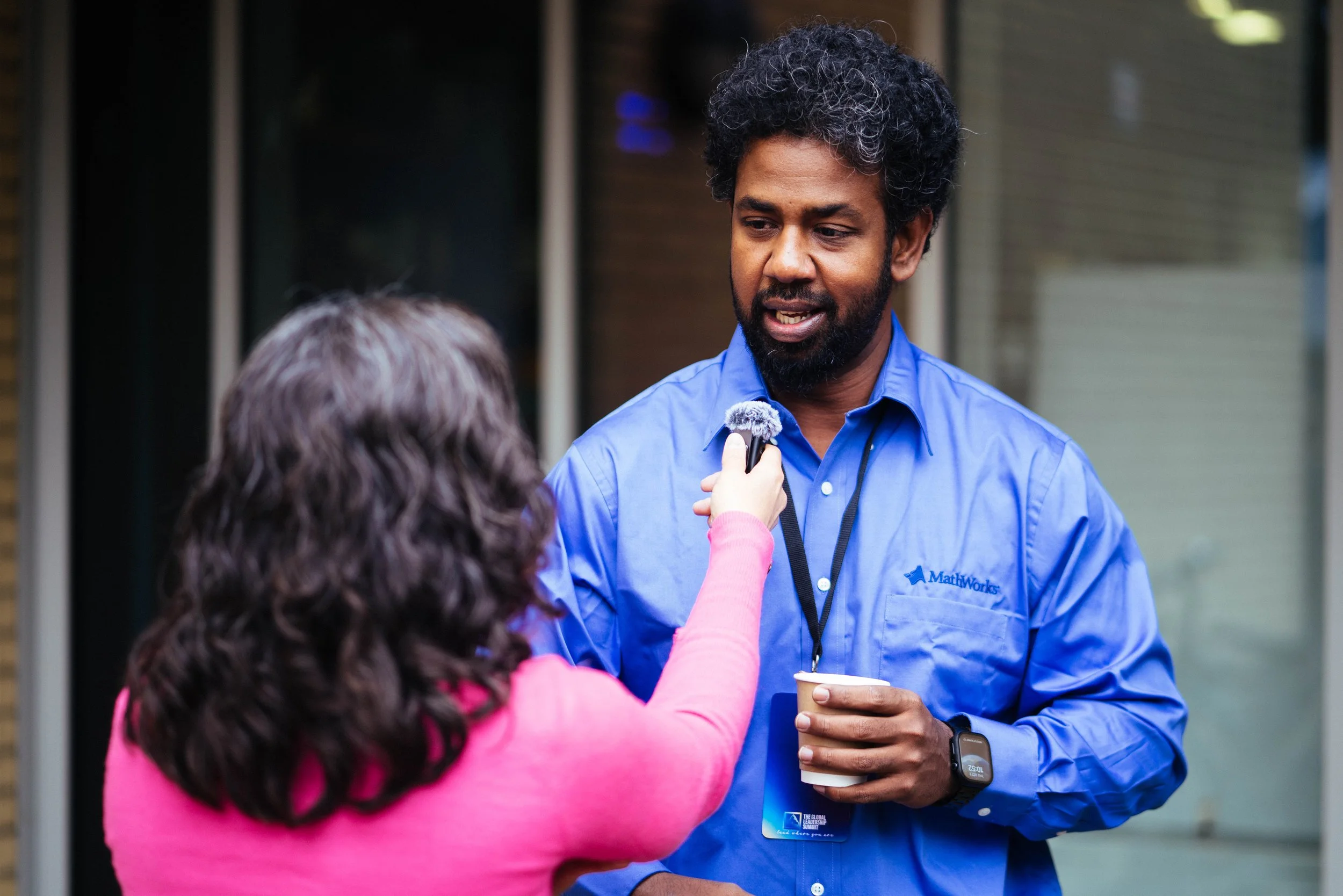 A man with curly black hair and a beard wearing a blue 'MathWorks' shirt is being interviewed by a woman holding a microphone. He is holding a cup and wearing a smartwatch, standing outdoors with a blurred background.