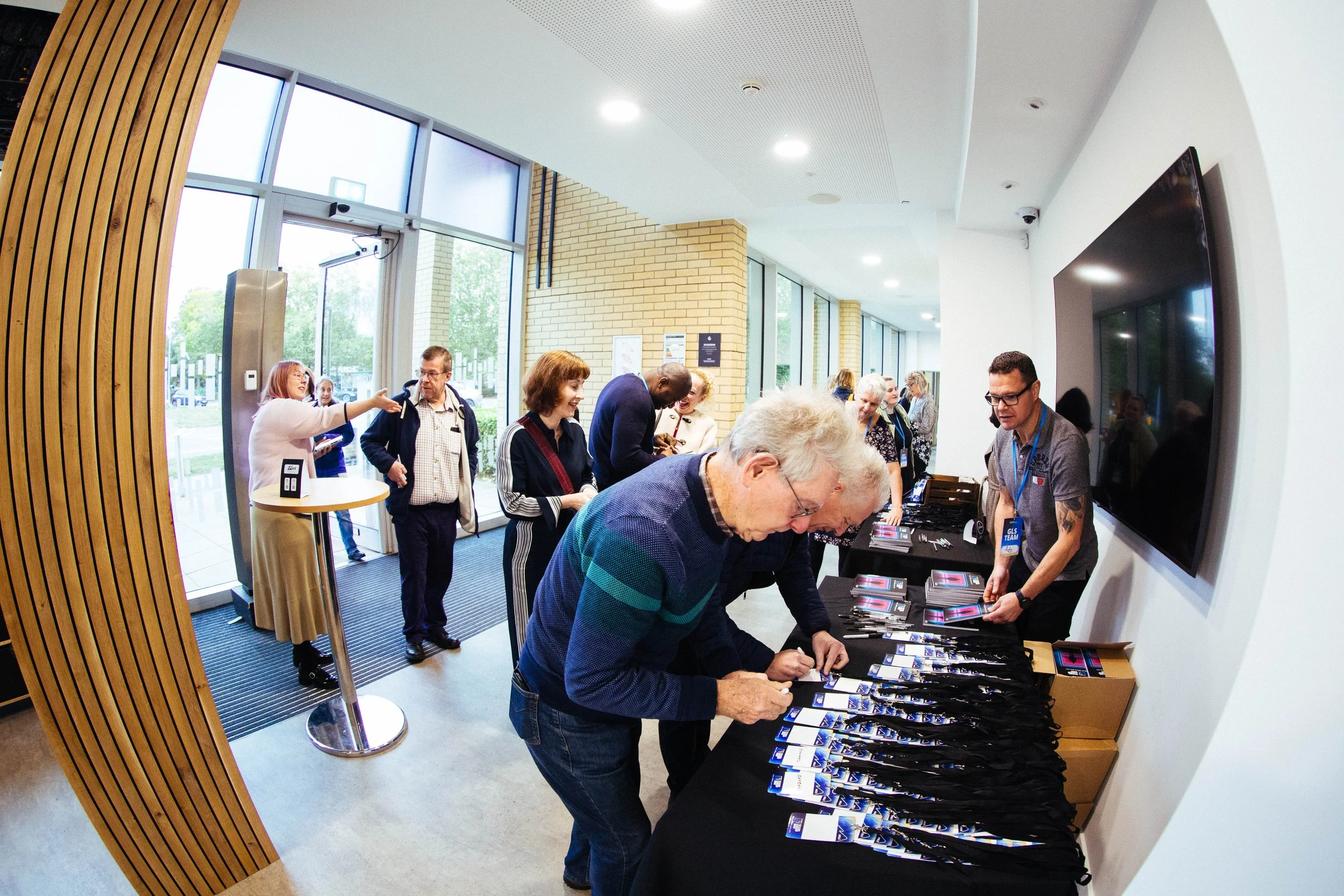 People gather around registration or information table at an indoor event, with large glass windows and wooden wall accents in the background.