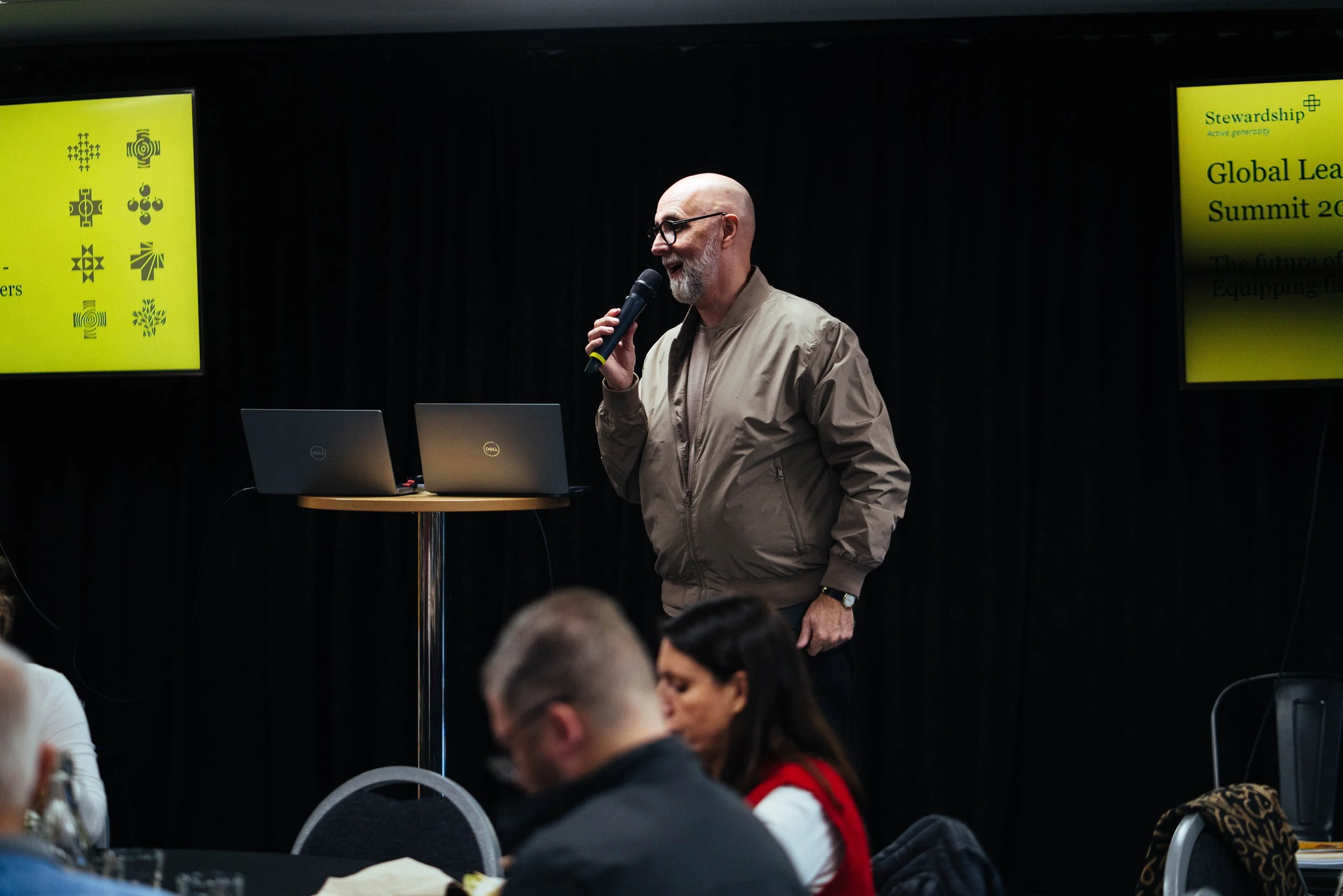 A man with glasses and a beard, holding a microphone, standing in front of two laptops during a presentation at a summit. There are yellow signage screens with logos and text about stewardship and the global leadership summit in the background.
