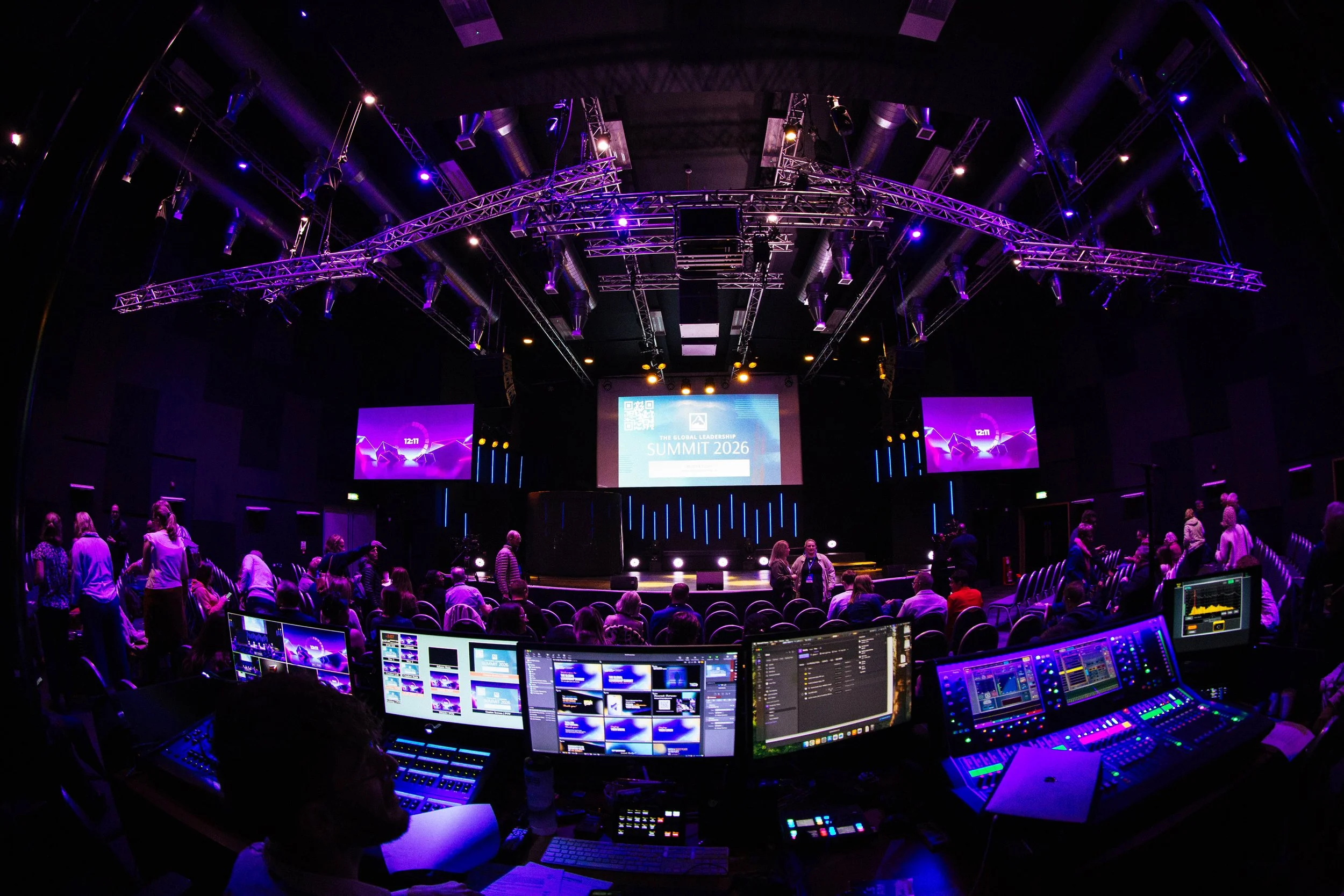 An event stage with purple and yellow lighting, a large screen reading 'The Global Leadership Summit 2026', and people seated and standing around in a dark auditorium with multiple screens and a technical control area in the foreground.
