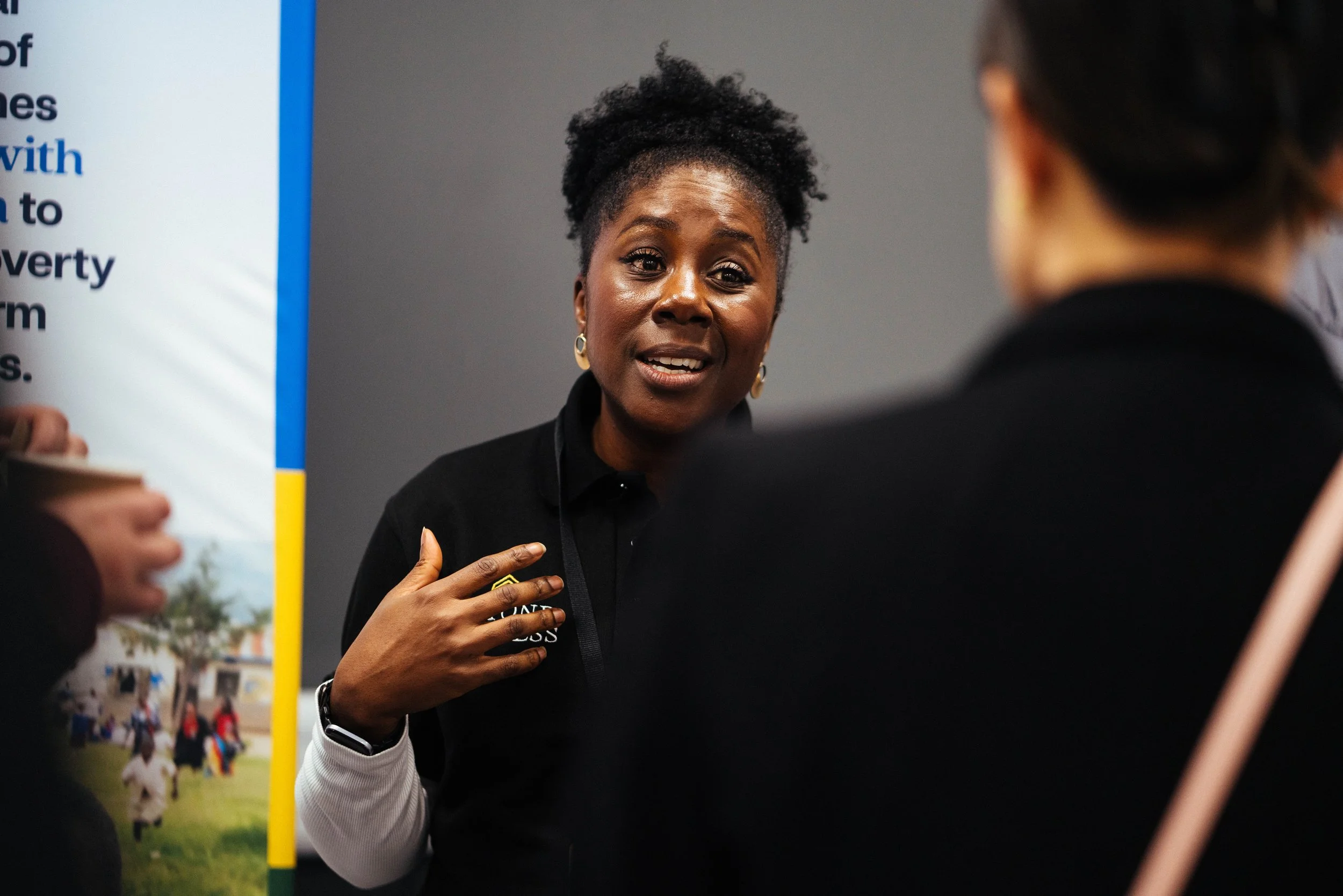 A woman with dark skin, curly ponytail, wearing a black shirt, is speaking to a person with short dark hair, who is partially visible. The woman is gesturing with her right hand and appears to be engaged in conversation. Behind her is a gray wall and