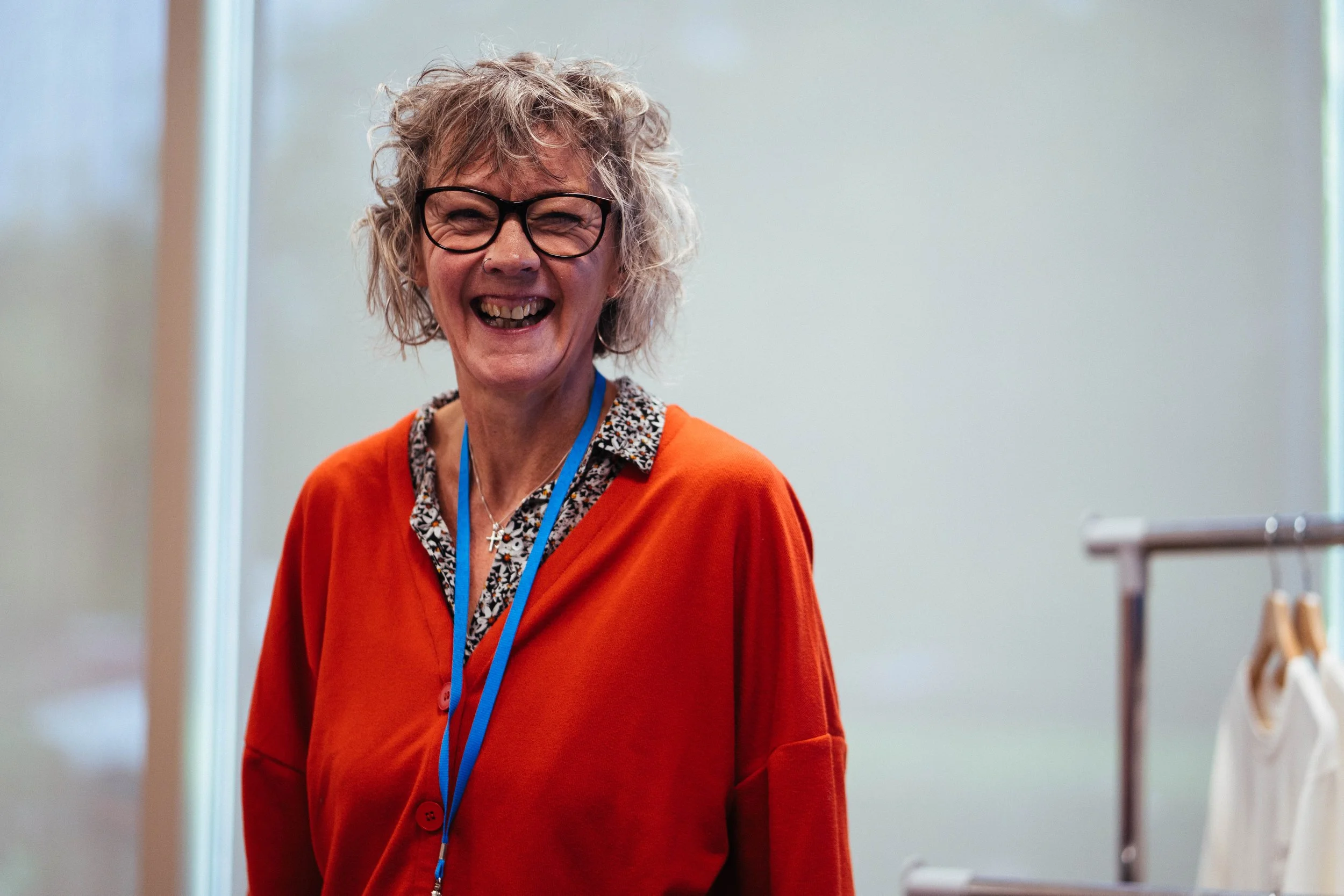 A woman with curly blonde hair, wearing glasses and a bright orange cardigan, smiling and laughing.