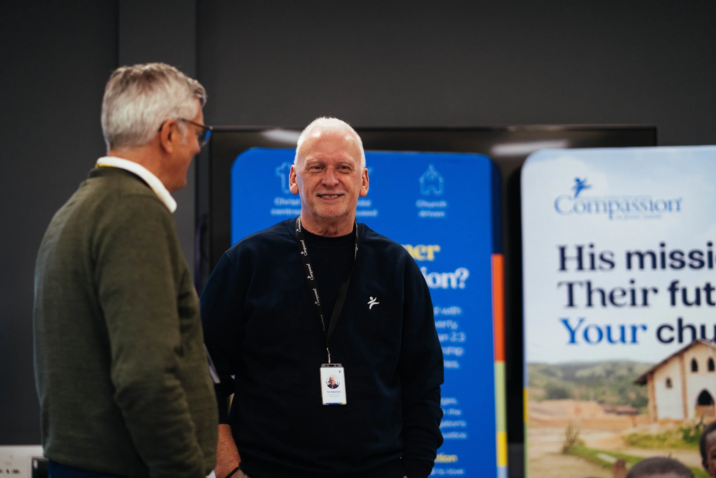 Two men conversing at an event with informational signs in the background, one with a name badge and lanyard, in an indoor setting.