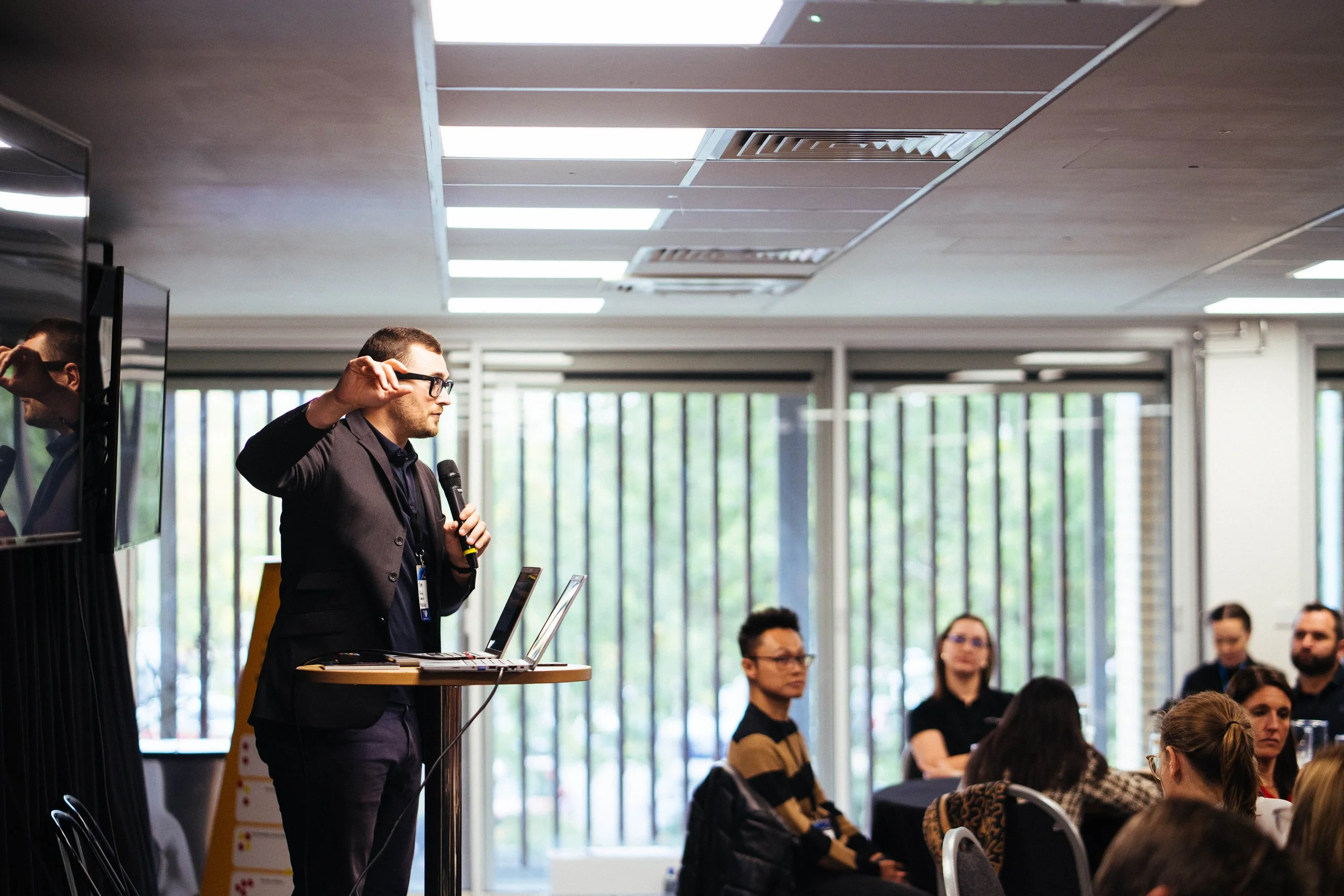 A man in a black blazer speaking into a microphone at a conference, standing at a presentation table with laptops, in a room with large windows and an audience seated and listening.