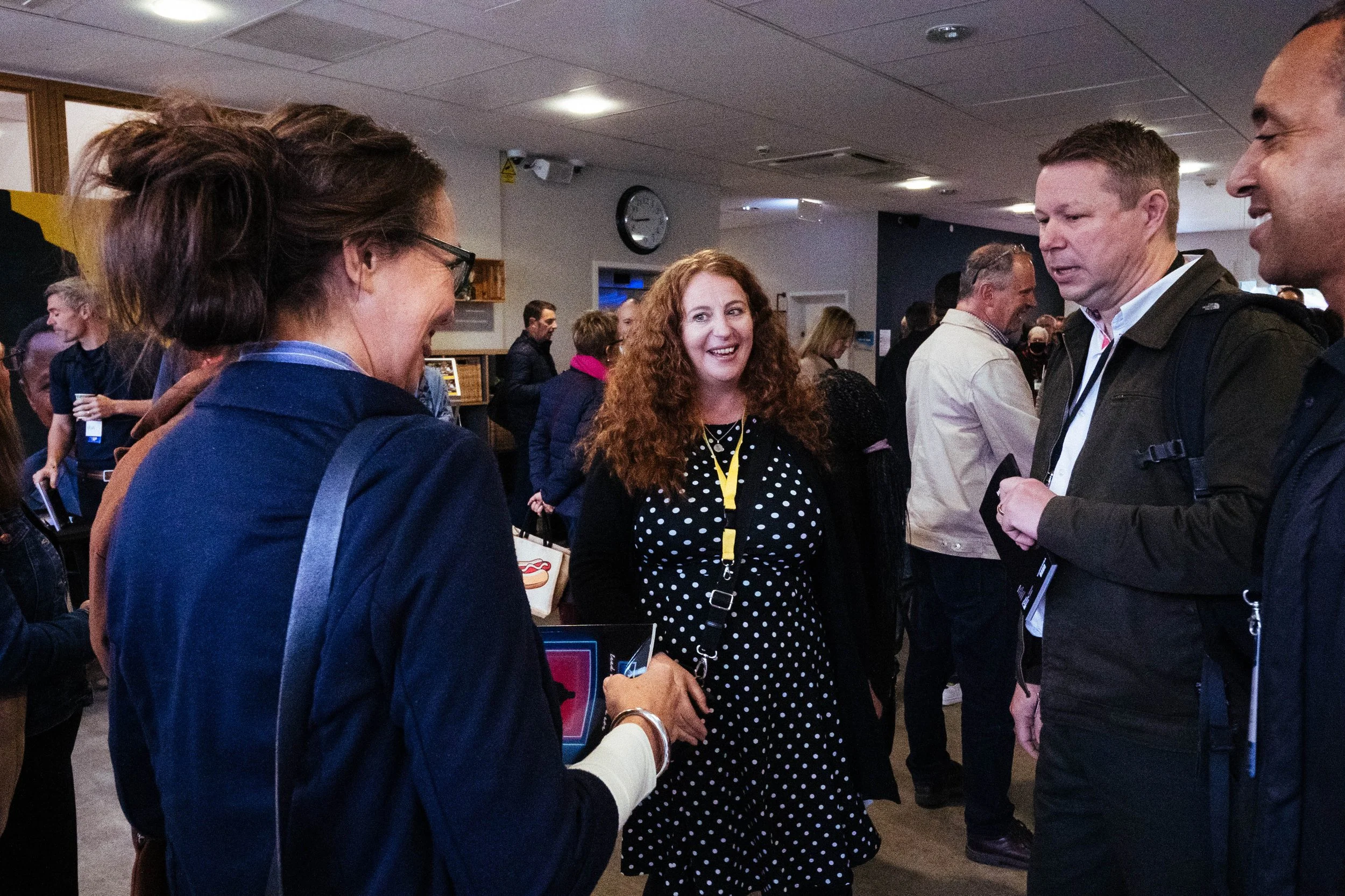 People interacting at an indoor event, with a woman with curly red hair wearing a black polka-dot dress talking to a man in a dark jacket. There are other people in the background, some holding drinks and engaging in conversations.