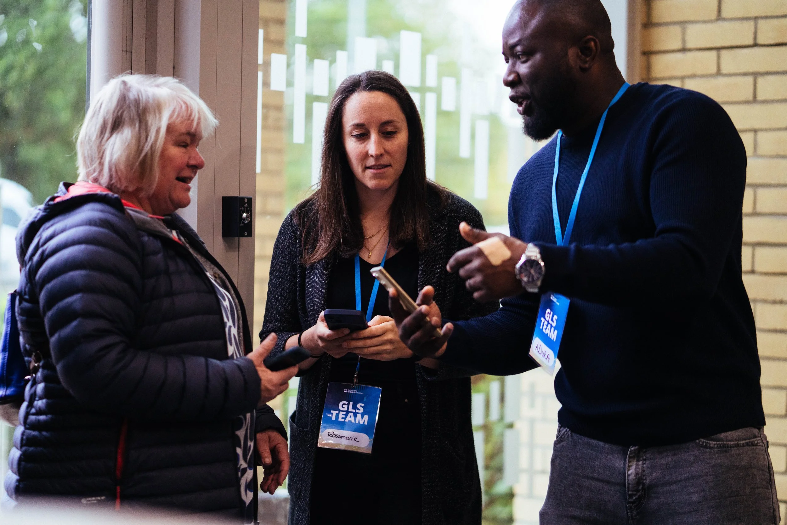 Three people engaging in conversation indoors near a large window. One woman with blonde hair wearing a dark jacket, a woman with brown hair wearing a business suit, and a man with a beard wearing a dark sweater. They are holding phones and wearing c