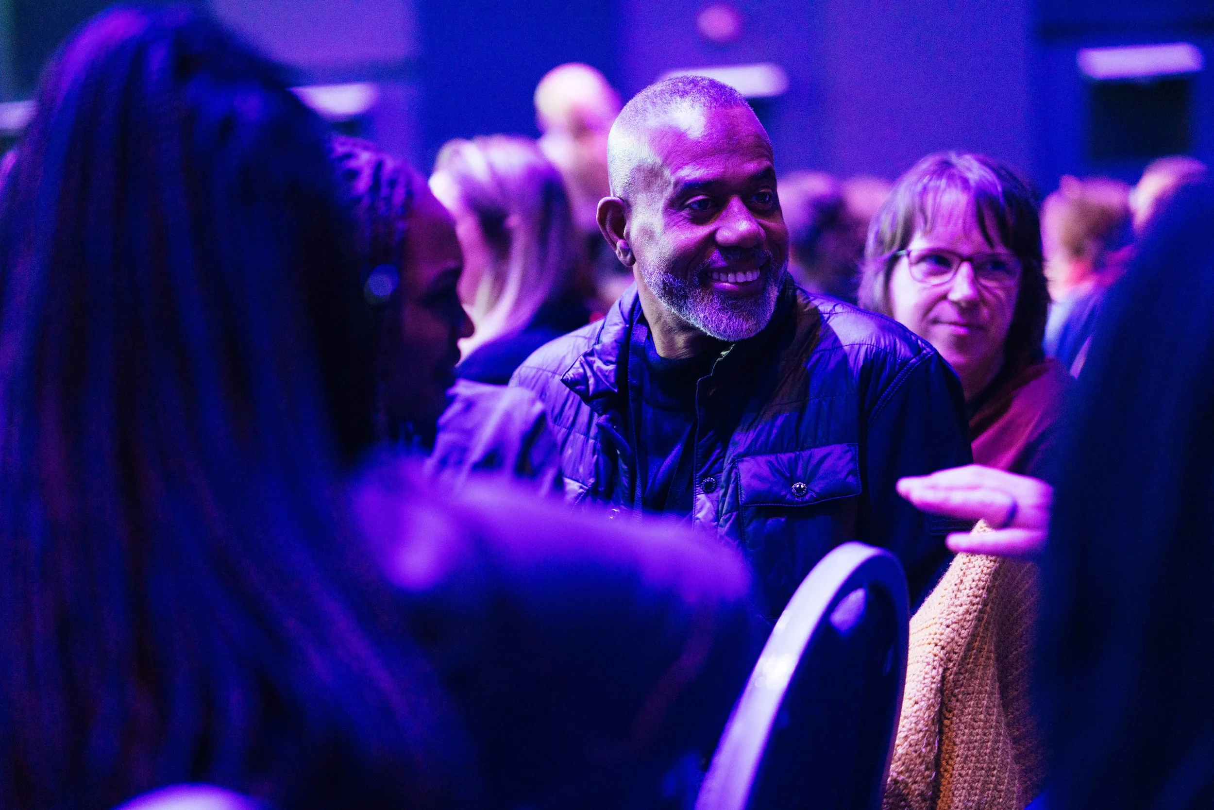People gathered in a dimly lit space with purple lighting, engaging in conversations, including a smiling man with a beard and a buzz cut wearing a black jacket.