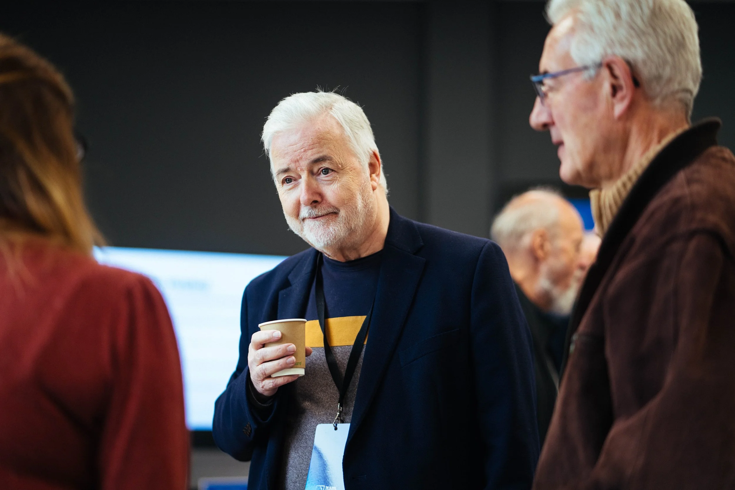 A man with white hair and a beard wearing a dark blazer and holding a paper coffee cup is engaged in conversation with two women at a professional event. The woman on the left has reddish-brown hair, and the woman on the right has short gray hair, an