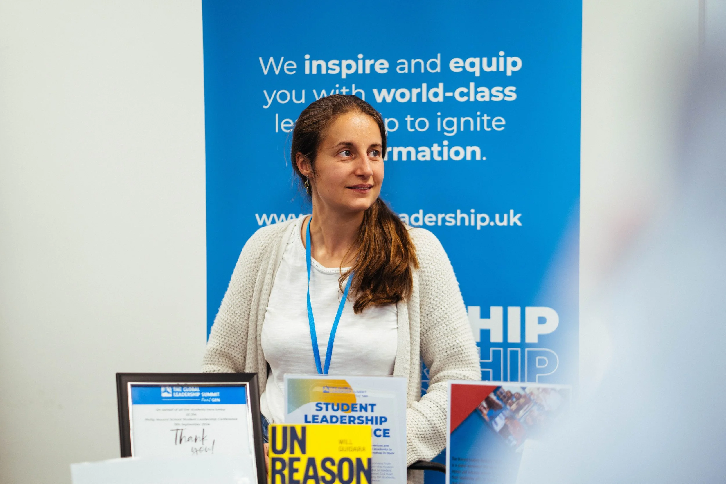 A smiling young woman with long brown hair and earrings stands in front of a blue banner that reads, "We inspire and equip you with world-class learning to ignite transformation." She is wearing a white shirt, a light cardigan, and a lanyard.