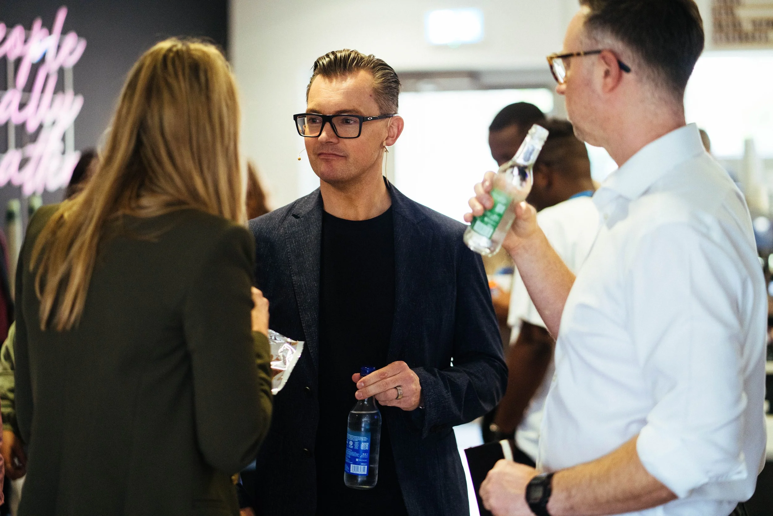 Three people conversing at a social gathering; one man with glasses and earrings, holding a water bottle, the woman with long hair has her back to the camera, and another man also wearing glasses and white shirt.