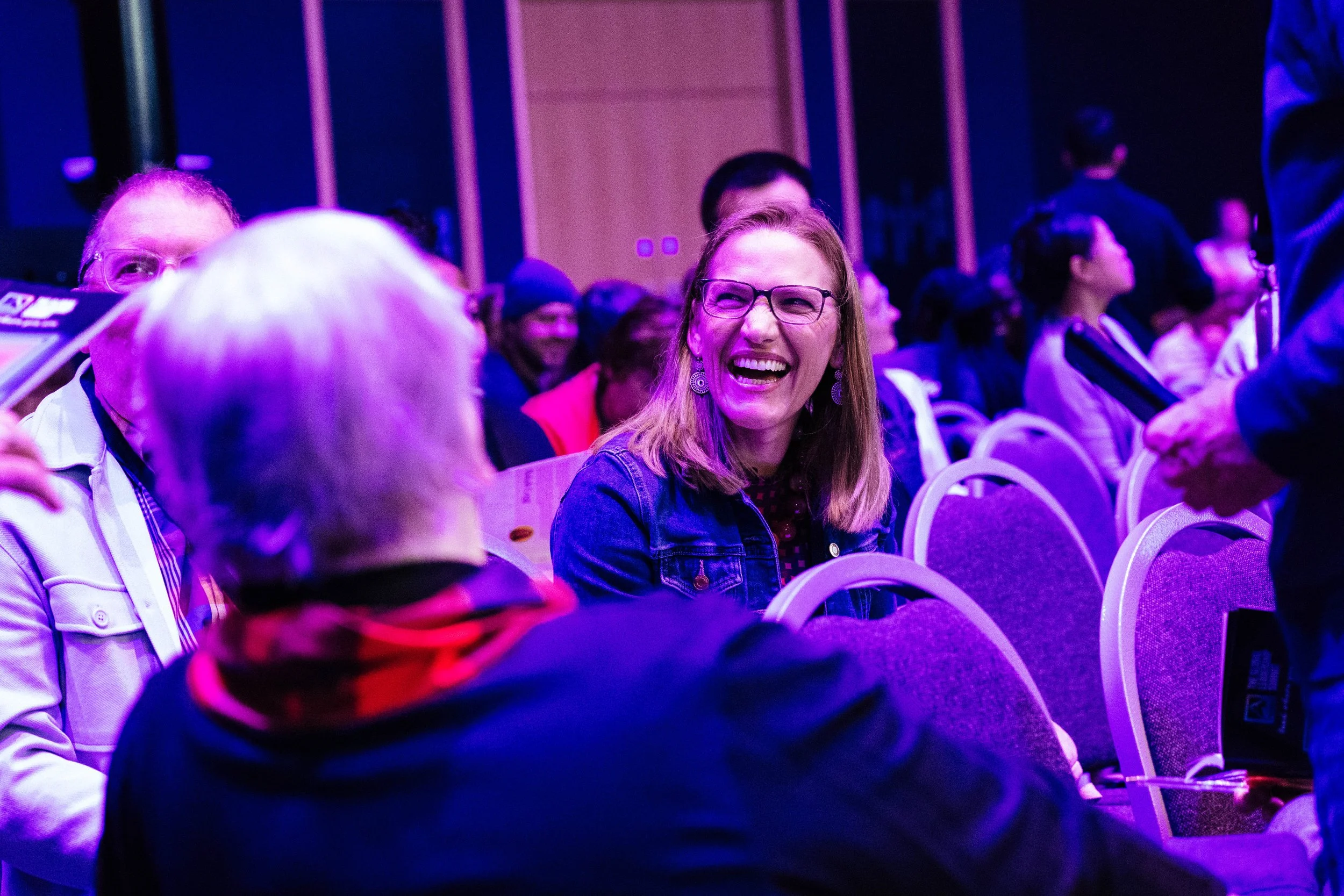 A group of people, including a woman with glasses and earrings, smiling and laughing, sitting in a dimly lit room with purple lighting, at what appears to be a social or conference event.