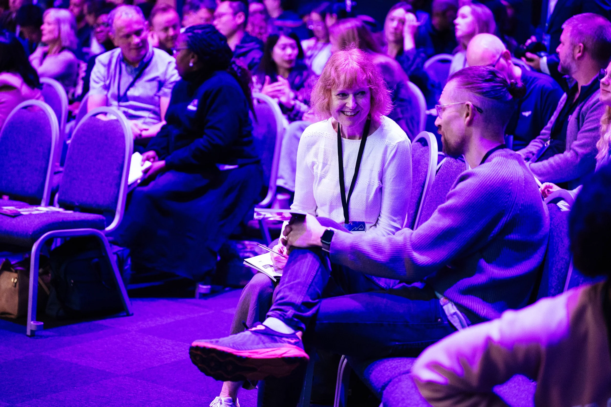 A diverse group of people sitting in chairs at a conference or event, with some engaged in conversation, and a woman with red hair smiling in the center, illuminated by purple lighting.