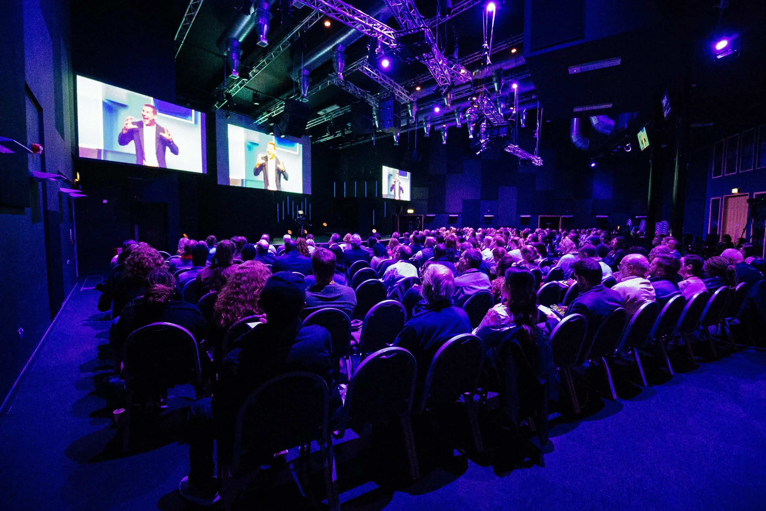 A large audience sitting in a darkened theater or conference hall, watching a presentation on a big screen. The presenter appears on the screen, gesturing with hands. The room is illuminated with purple and blue stage lights, and the ceiling has a me