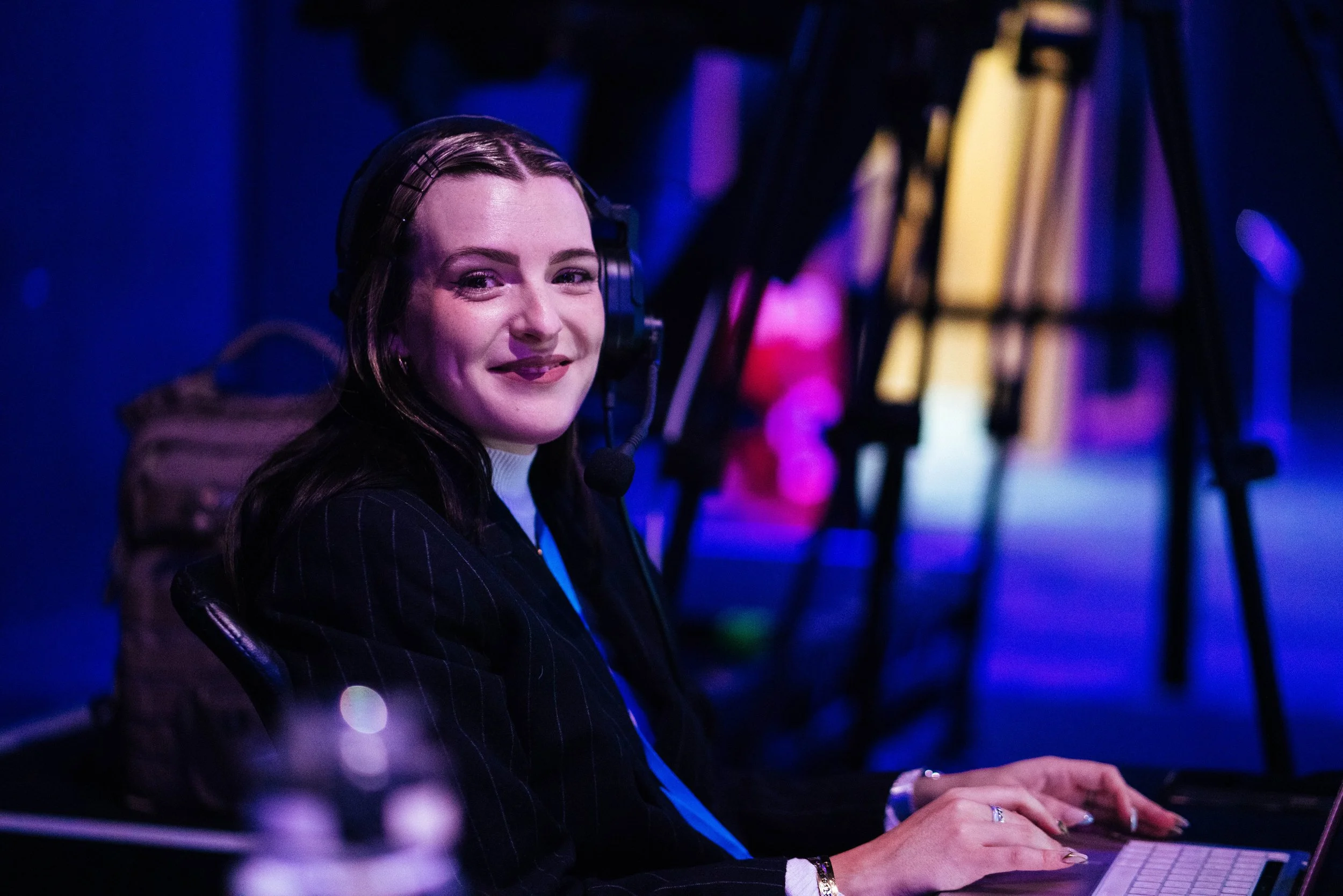 A woman with long dark hair, wearing a pinstripe suit, is smiling while playing a keyboard in a dark, colorful setting with purple and yellow lights.
