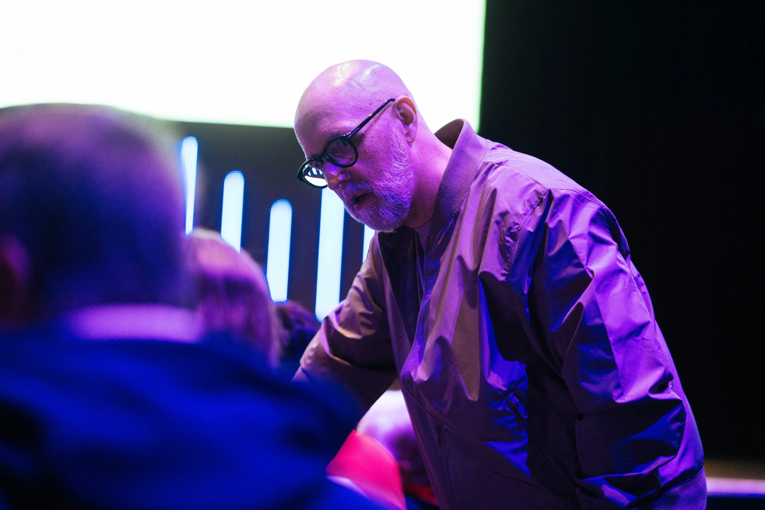 A man with glasses, a beard, and a bald head, wearing a tan jacket, leans forward in a dark setting with purple and green lighting, seemingly focused on an activity or object in front of him.