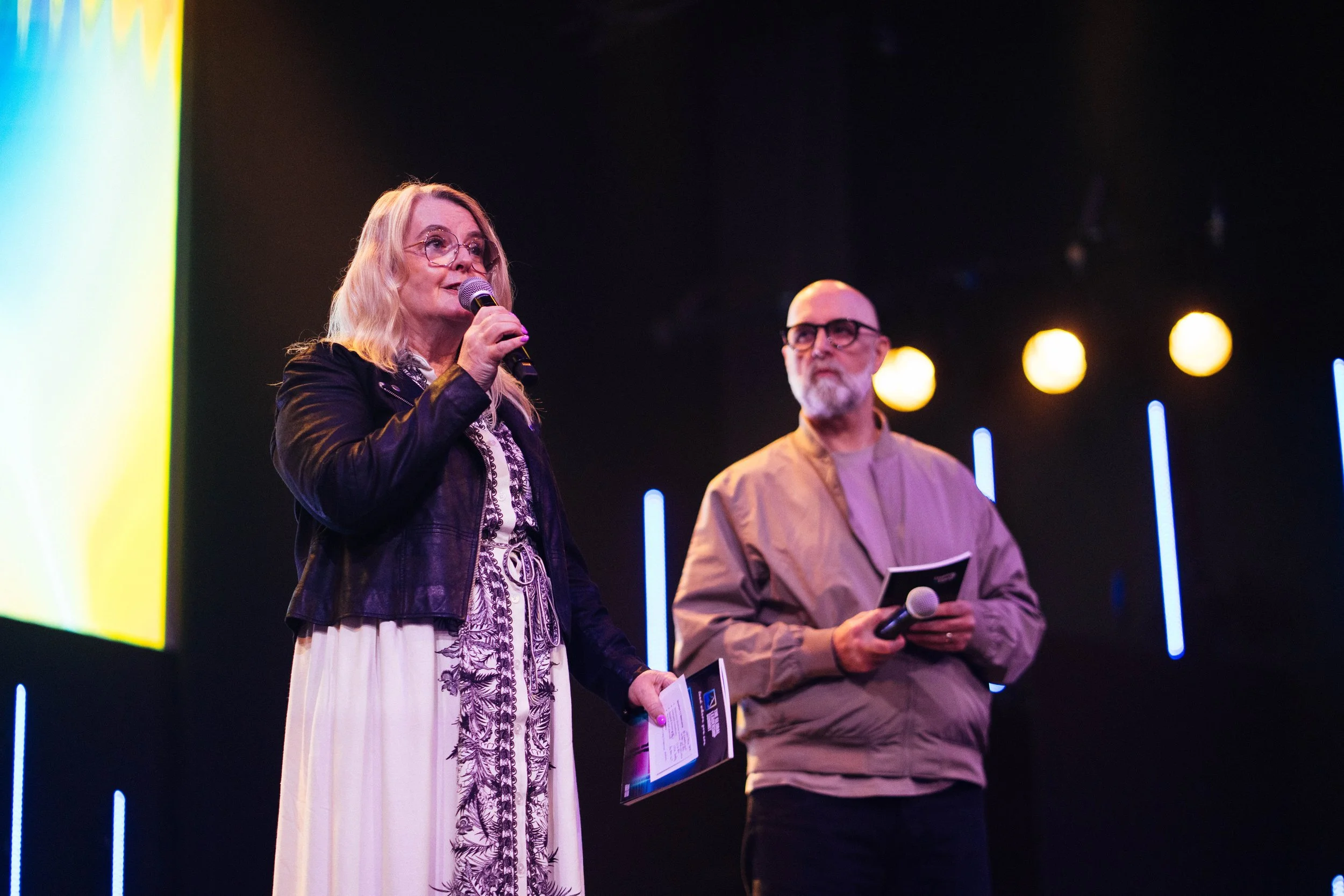A woman with blonde hair and glasses speaking into a microphone, wearing a black leather jacket and a white dress with a black embroidered pattern. Next to her, a man with a bald head, glasses, and a gray beard is holding a microphone and a booklet. 