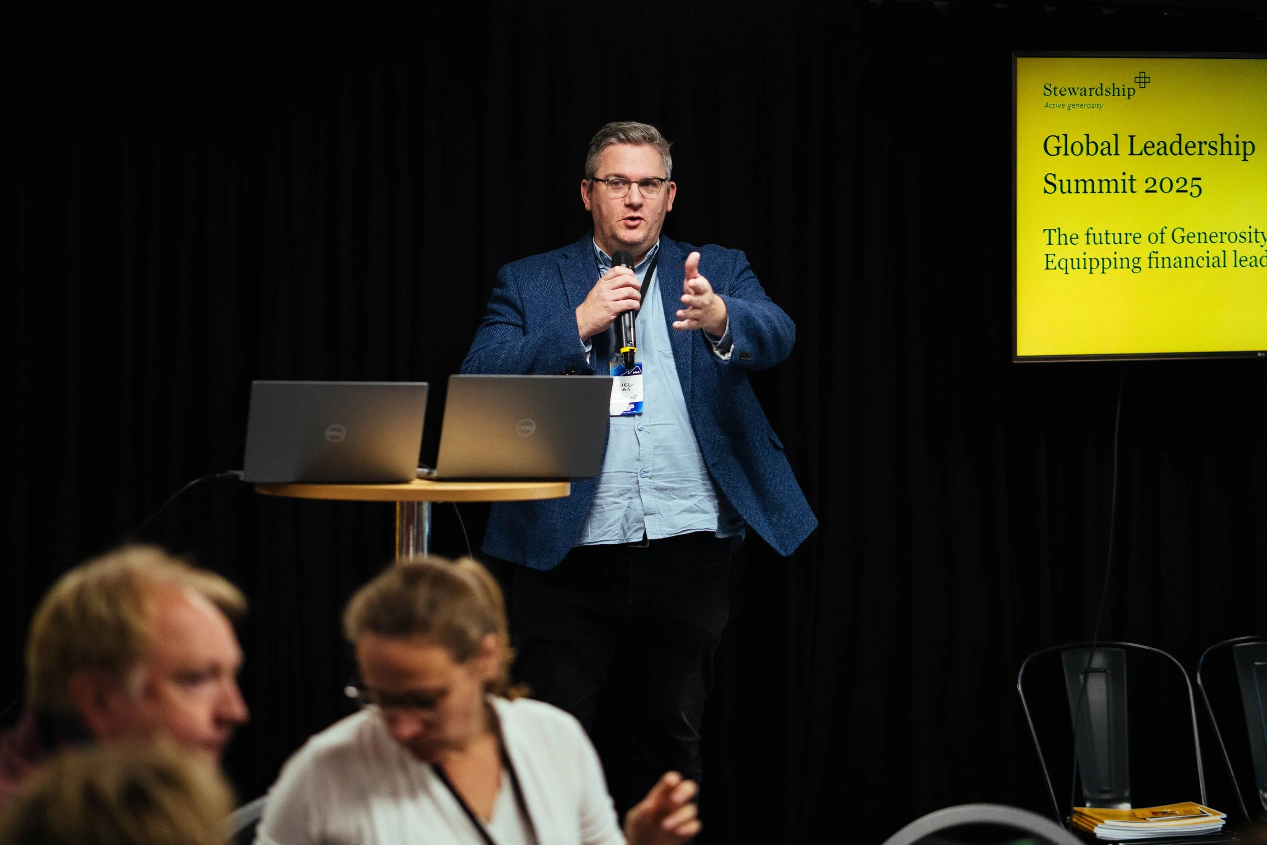 A man in glasses and a blue blazer is speaking into a microphone during a conference. Behind him, there is a yellow screen with text about a leadership summit.