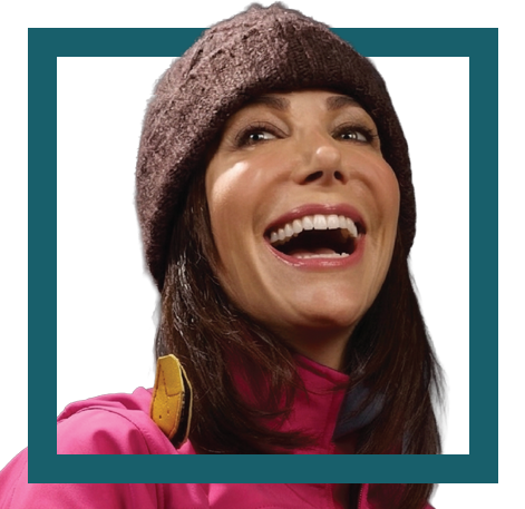 Close-up of a woman smiling, wearing a brown beanie and pink jacket, with a backpack strap visible.